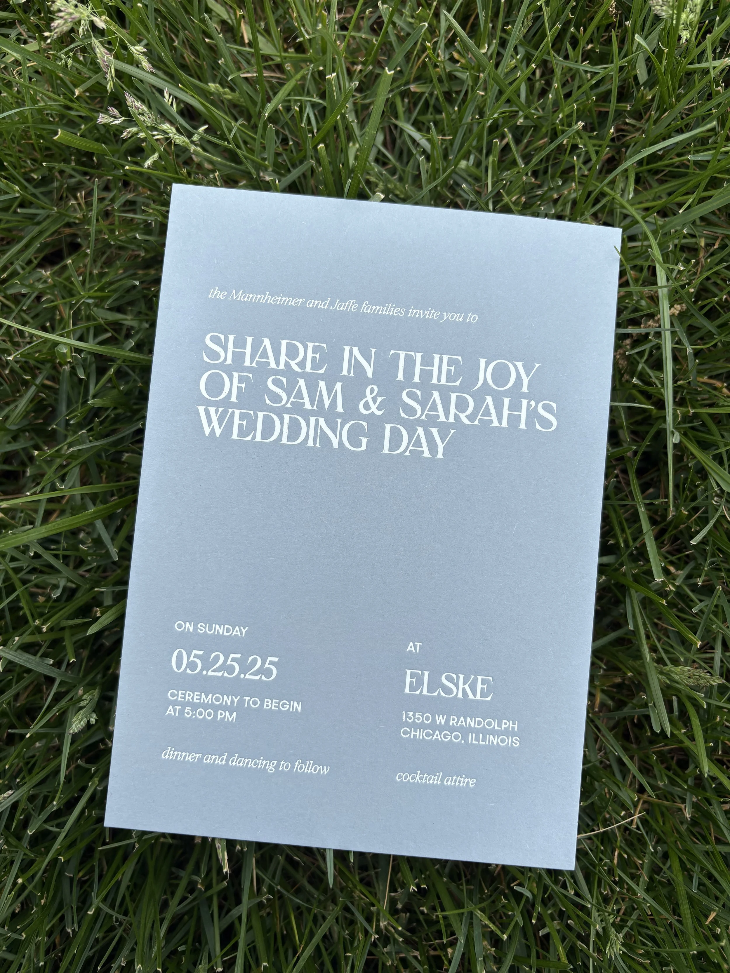 A light blue wedding invitation card on green grass, with details about Sam and Sarah's wedding, including date, time, location, and dress code.