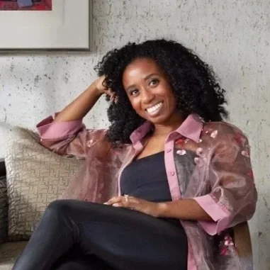 A woman with curly black hair smiling and sitting on a couch, wearing a pink and brown jacket over a black top.