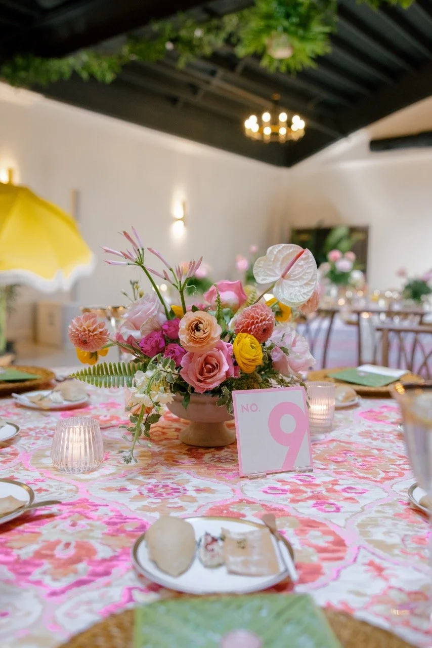A table decorated with a pink floral tablecloth and a pink and white floral centerpiece, surrounded by place settings with plates and utensils, pink candles, and a table number card with a pink number nine, in a bright, elegant event space.