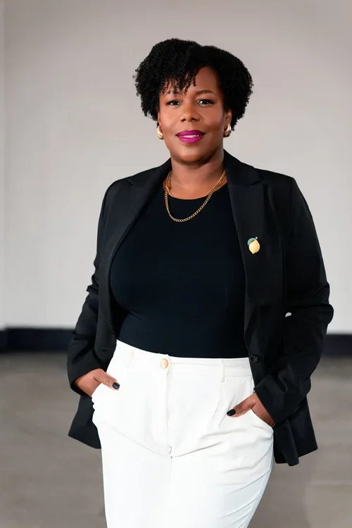 A confident Black woman with short curly hair wearing a black blazer over a black top, white trousers, and gold jewelry, standing in an indoor setting with a neutral background.