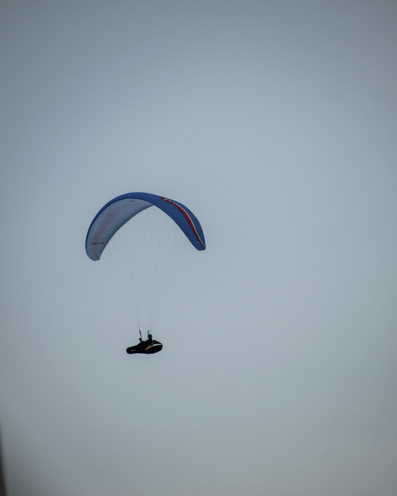 A person paragliding with a blue, red, and white parachute in an overcast sky.