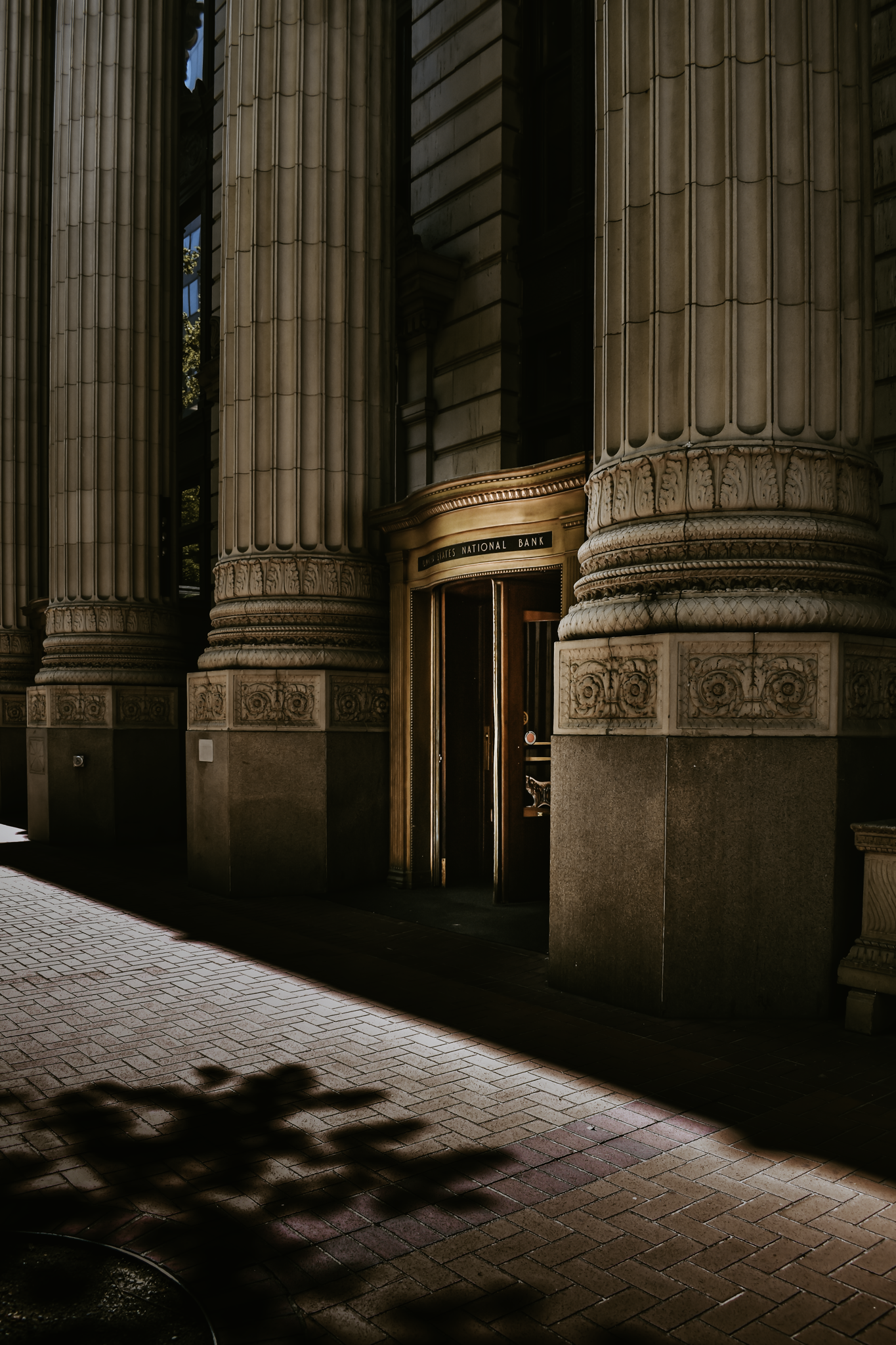 Exterior of a historic bank building with large stone columns and a revolving door entrance, with shadows cast on the brick sidewalk.