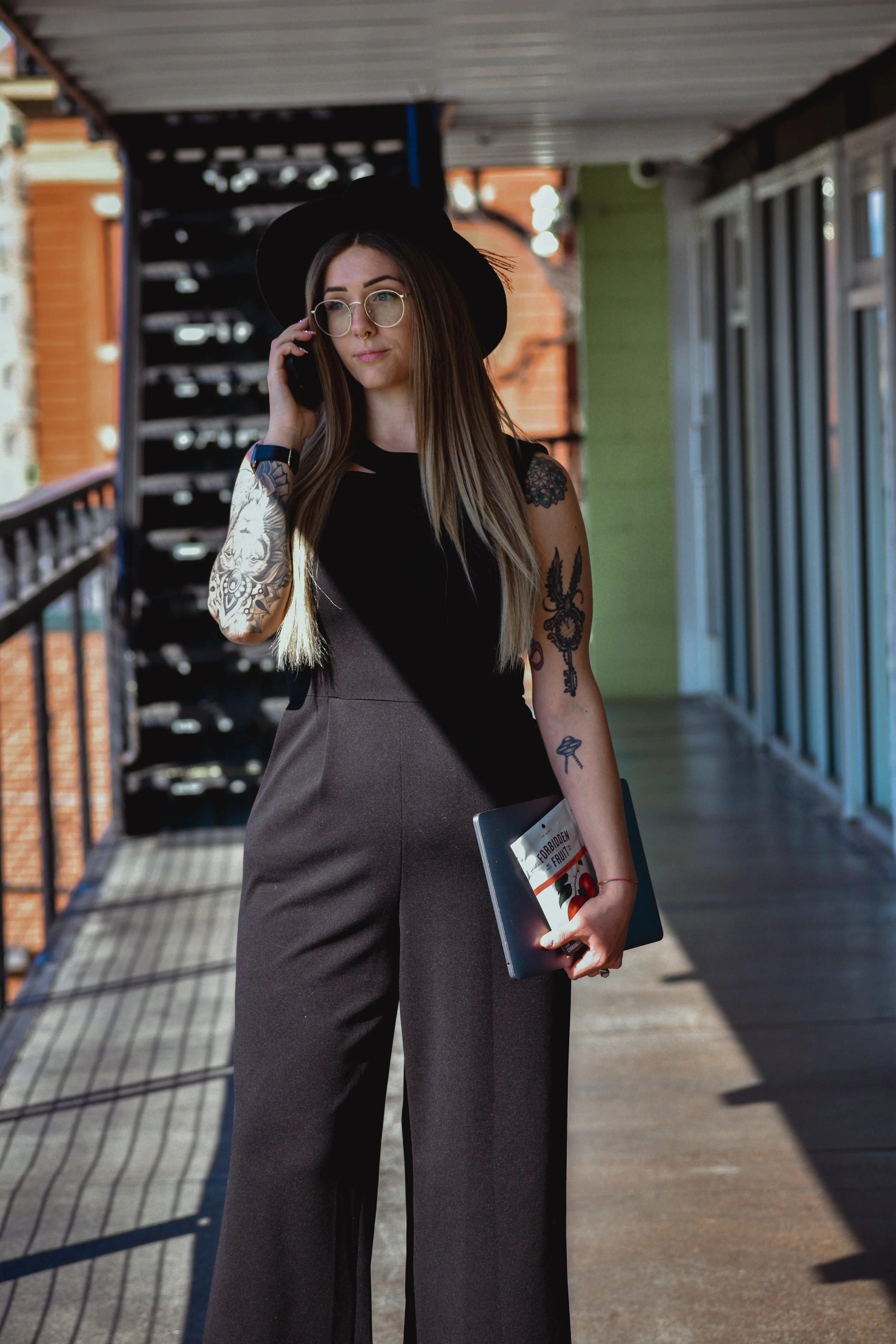 Young woman in black hat and outfit standing on balcony, talking on phone, holding a book titled 'Forbidden Fruit' with a fruit illustration, tattoos on her arms, sunglasses, long brown hair.