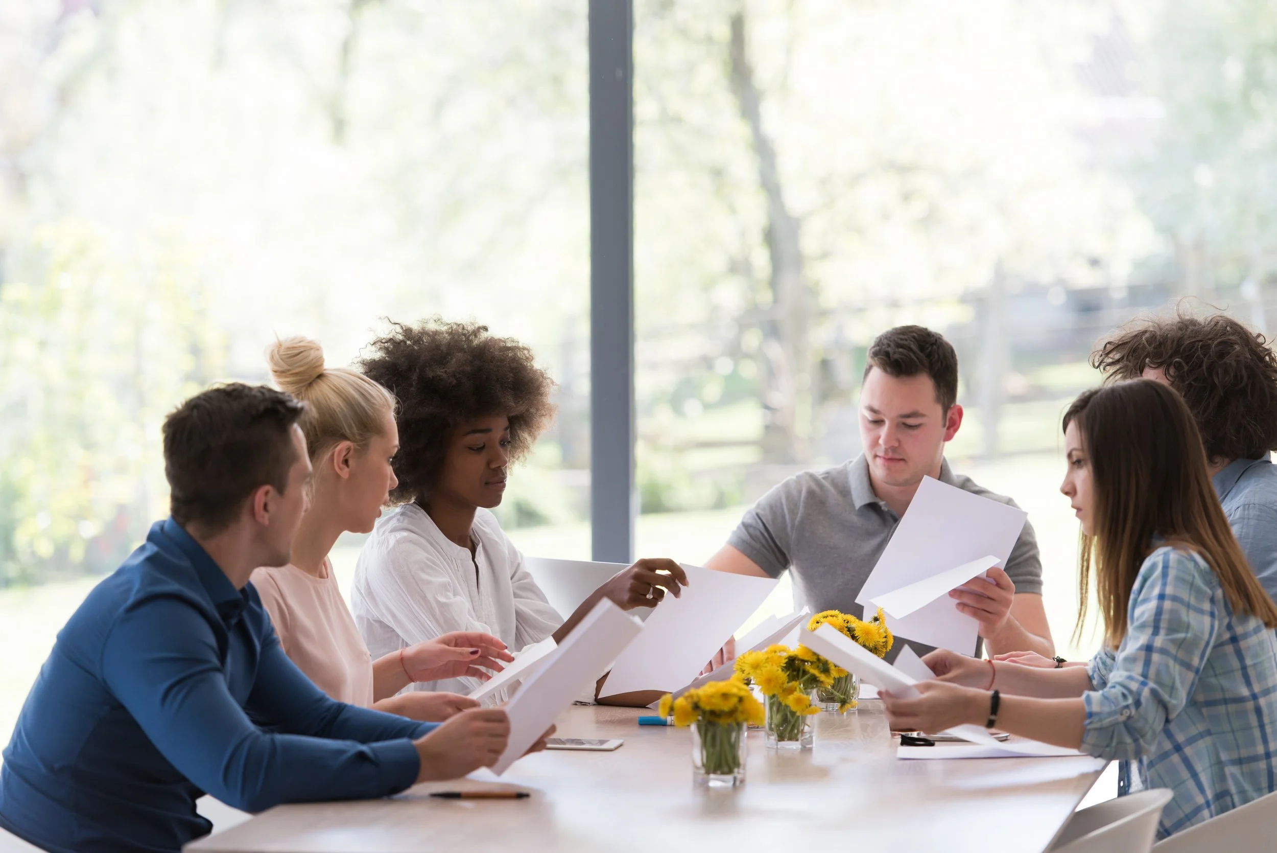 group of people meeting around a table with flowers in glasses, holding papers