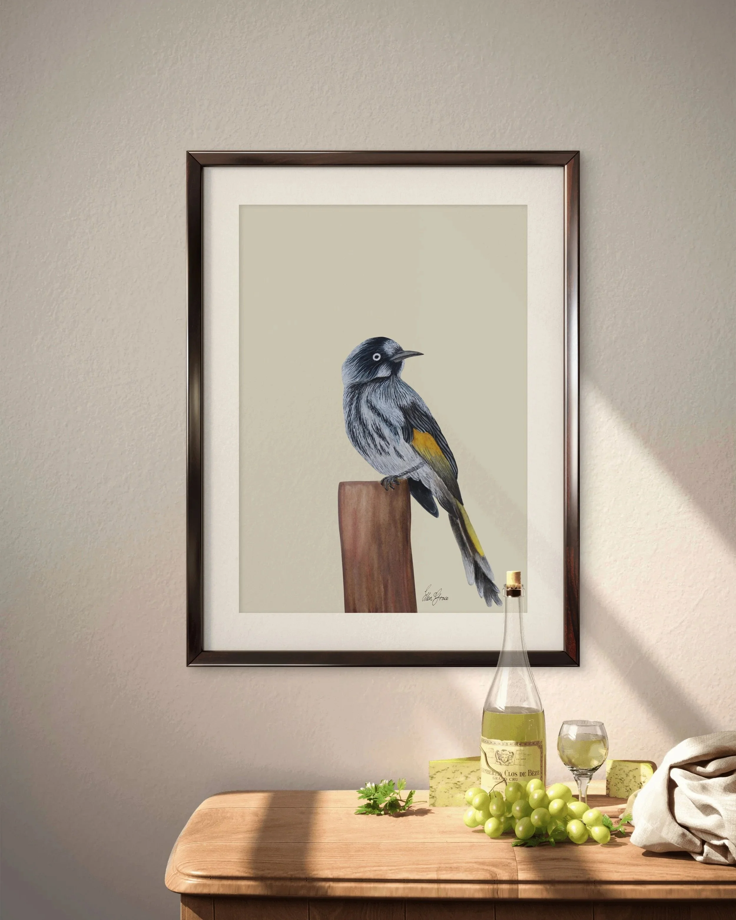 Framed artwork of a New Holland Honeyeater perched on a wooden post, hanging on a beige wall above a wooden table set with white wine, grapes, cheese, and a folded cloth.