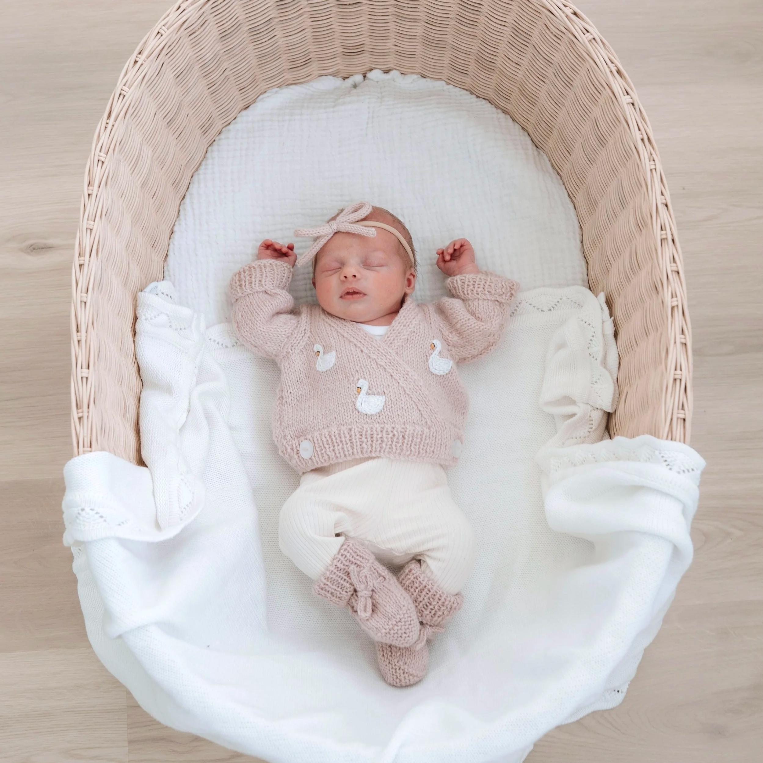 A sleeping baby dressed in pink embroidered knit cardigan and white clothing, with a pink bow headband, lying in a natural wood crib with white blanket and bedding.