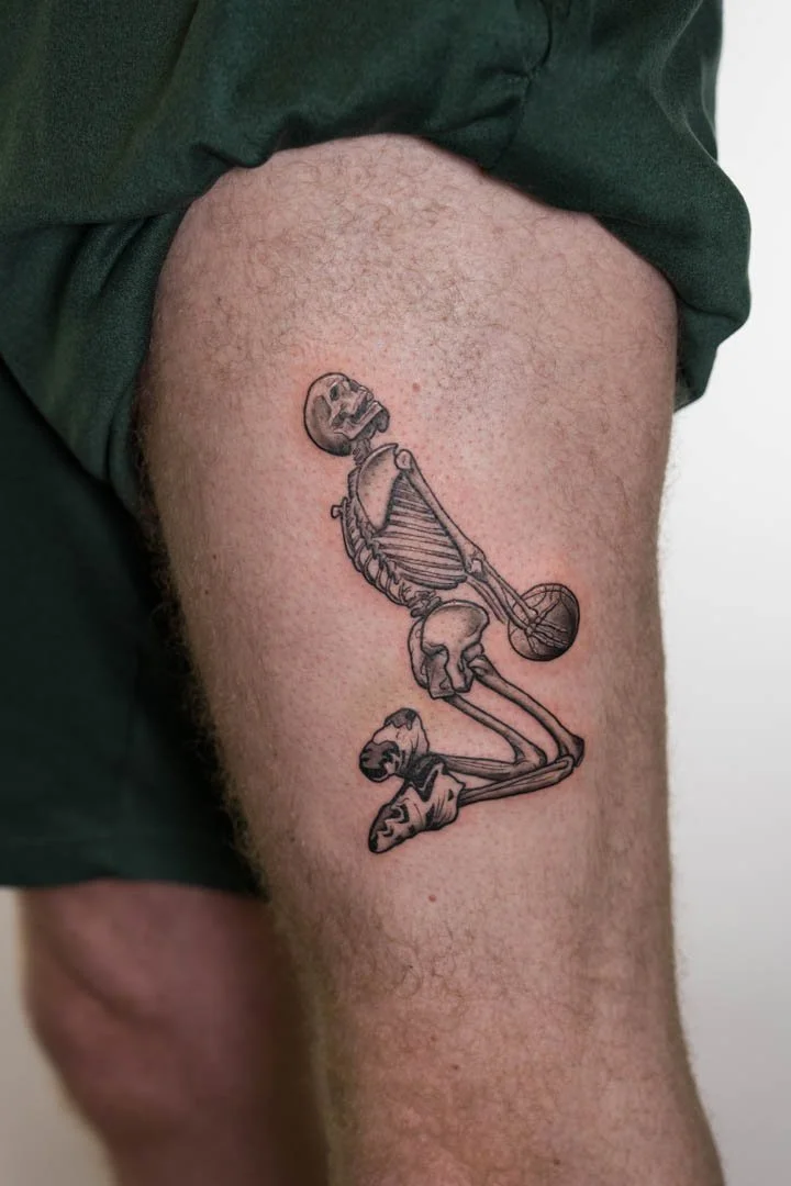 Photograph of a tattoo of and iconic Kobe Bryant dunk as a skeleton. Skeleton is holding a basketball and wearing Kobe Byrant signature Adidas sneakers. The placement of the tattoo out on the outer left thigh. The style the tattoo is graphic black an