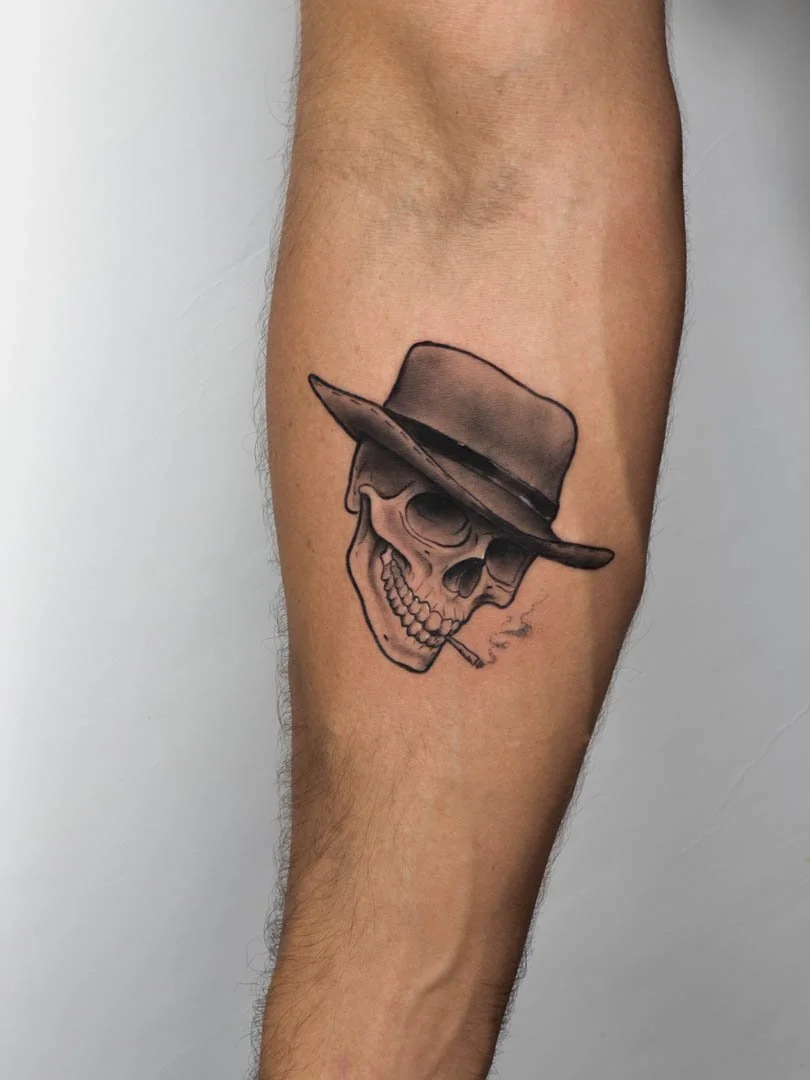Tattoo of a cowboy skull. Skull is wearing a cowboy hat and has a cigarette in it’s mouth. The tattoo is positioned on the inner right forearm. Style is soft black and grey