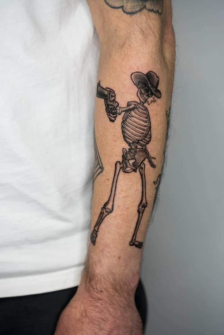 Cowboy skeleton holding a revolver. Placement is on the outer left forearm. Style is illustrative black and grey.