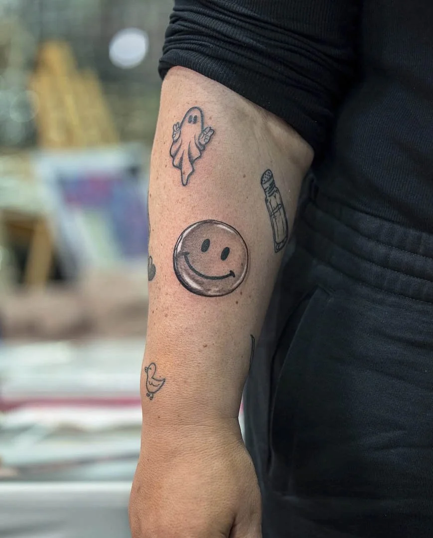 Chrome smiley face tattoo placed on the front of the right forearm. The tattoo is black and grey realism / chrome effect.