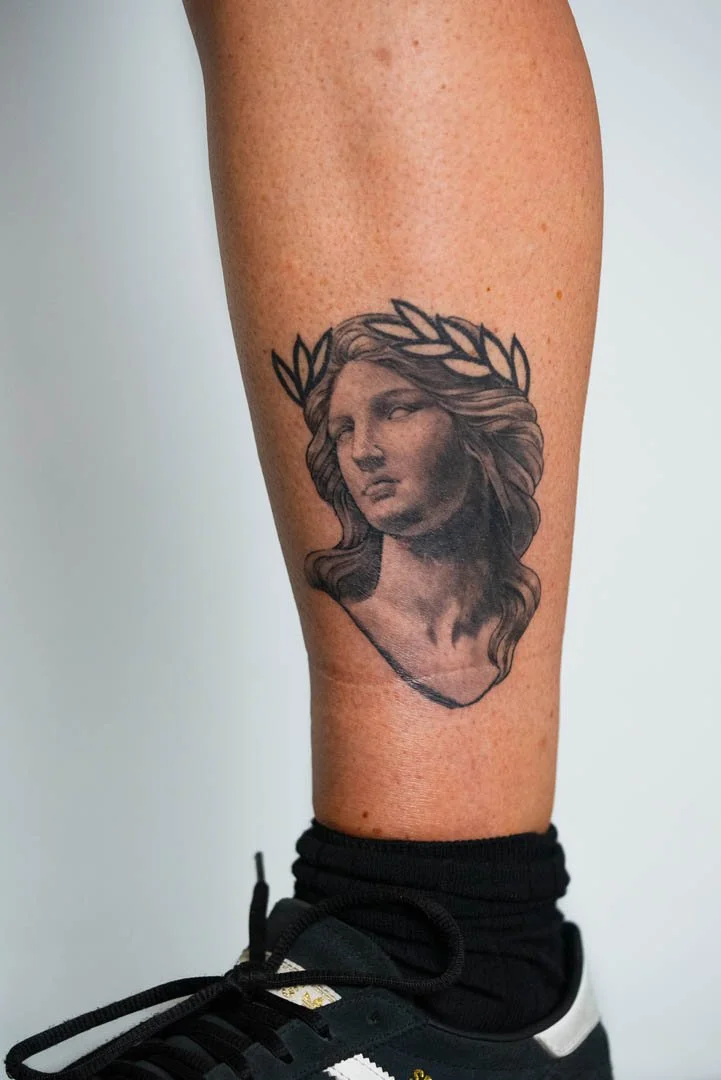 Greek statue bust of the goddess Nike wearing a laurel wreath. Placement is outer left calf. Style is soft black and grey with bold graphic elements