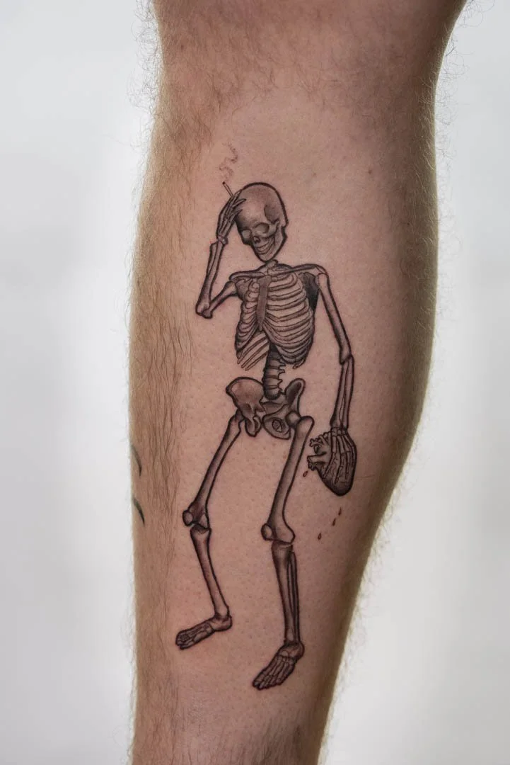 Tattoo of a skeleton with one hand on its head and the other hand holding a bleeding heart. The placement is on the inner left calf. The style of the tattoo is graphic black and graphic.