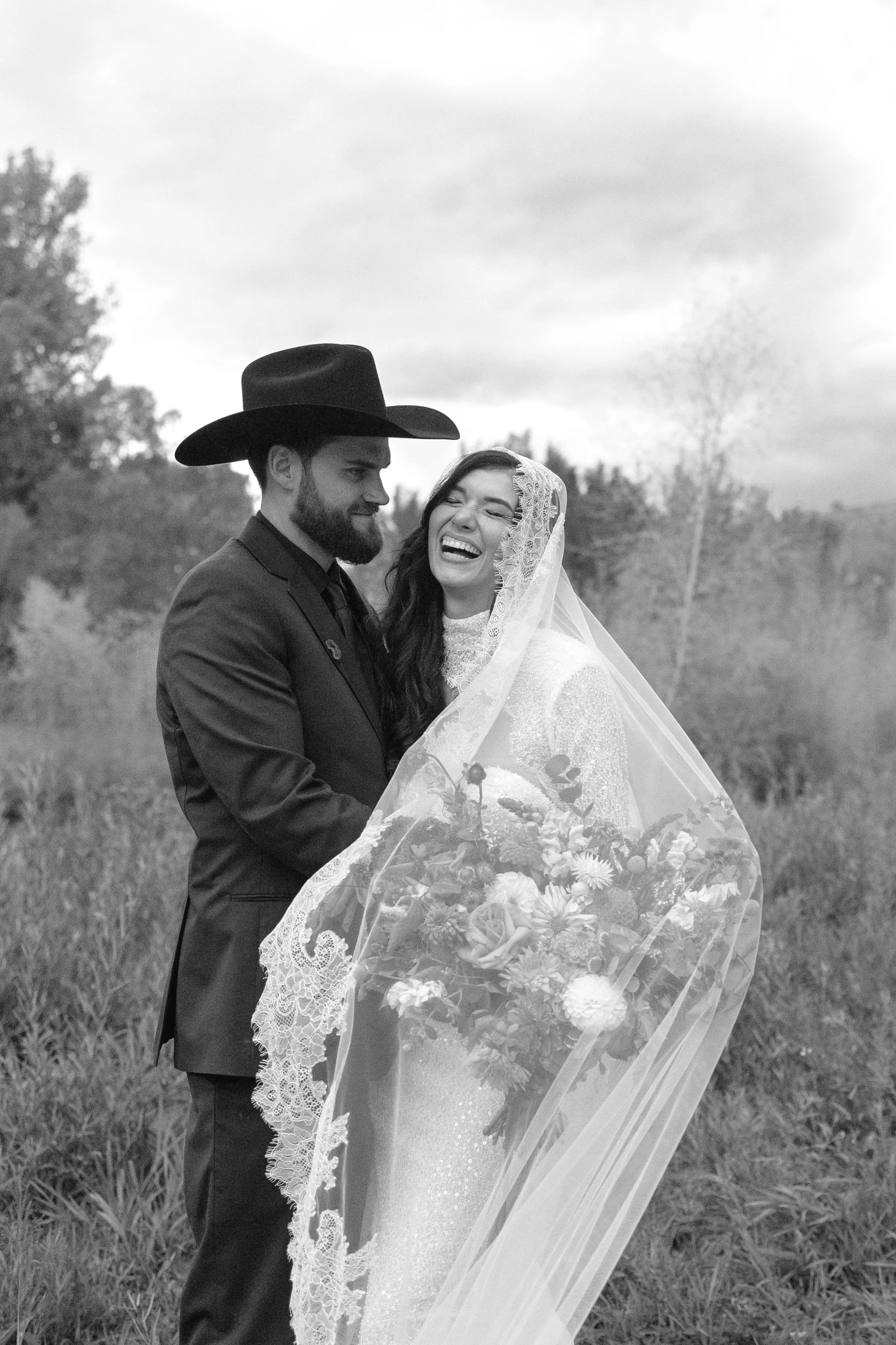 A black and white photo of a bride and groom outdoors, with the groom wearing a cowboy hat and the bride laughing, holding a bouquet, and wearing a lace veil.