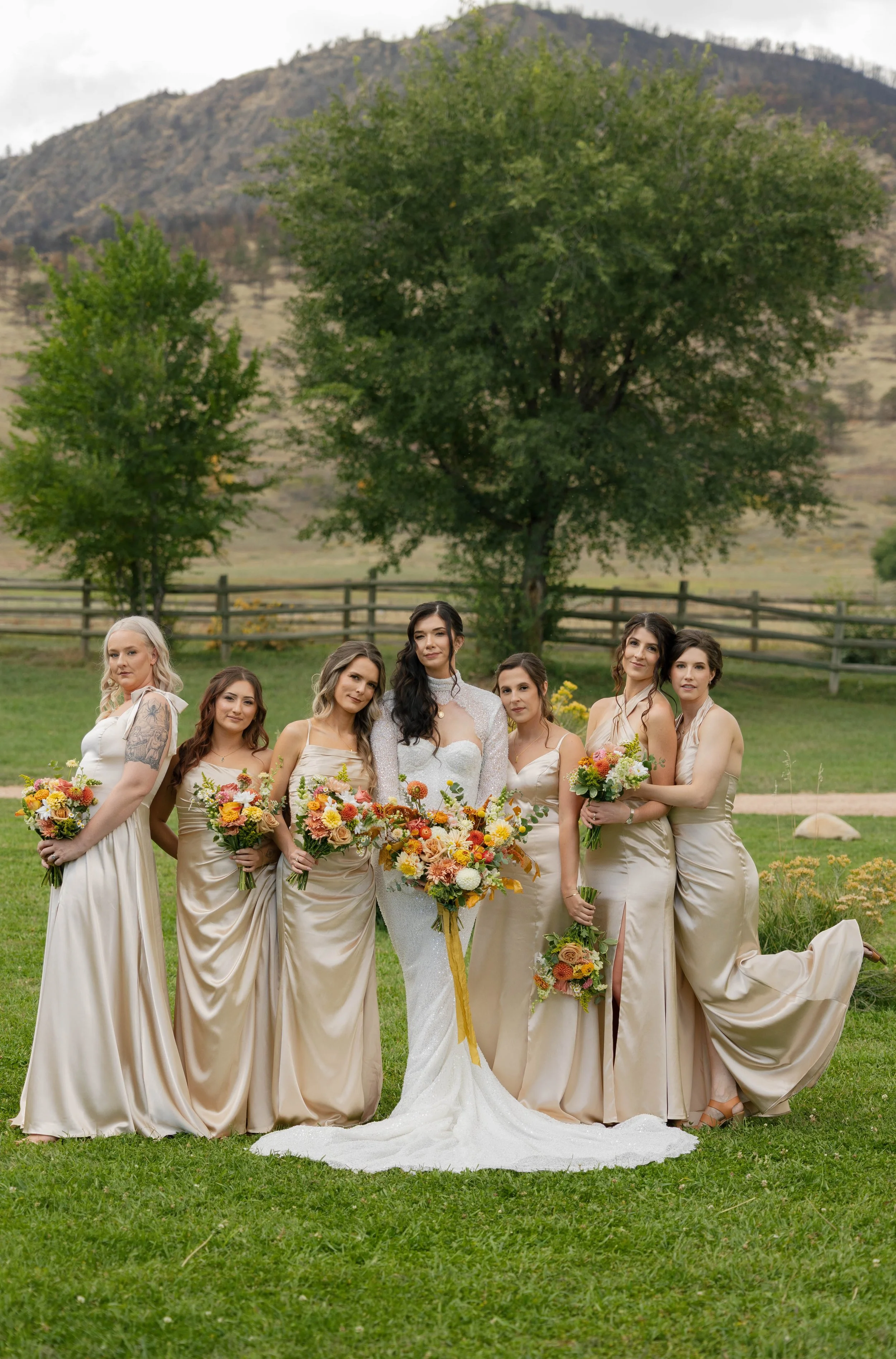 Group of seven women in formal dresses, standing outdoors on green grass with trees and hills in the background, holding bouquets of flowers.
