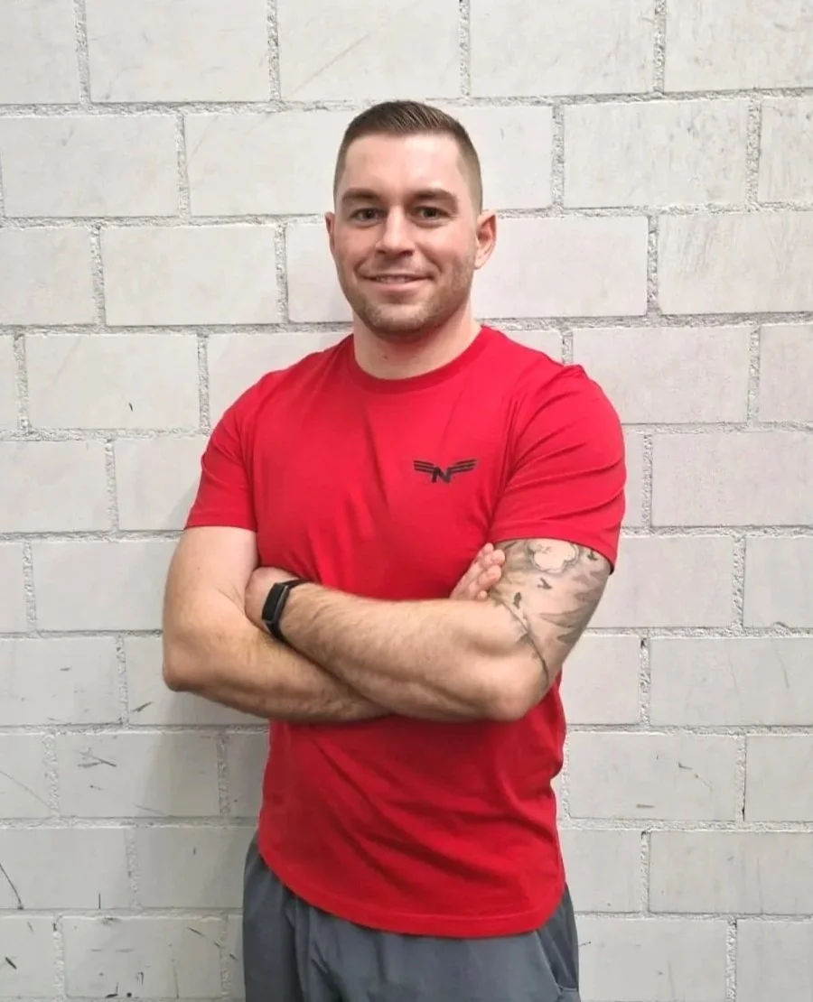 Ein Mann steht vor einer weißen Backsteinmauer, trägt ein rotes T-Shirt mit einem schwarzen Logo und hat Tattoos am linken Arm. Er lächelt und verschränkt die Arme.