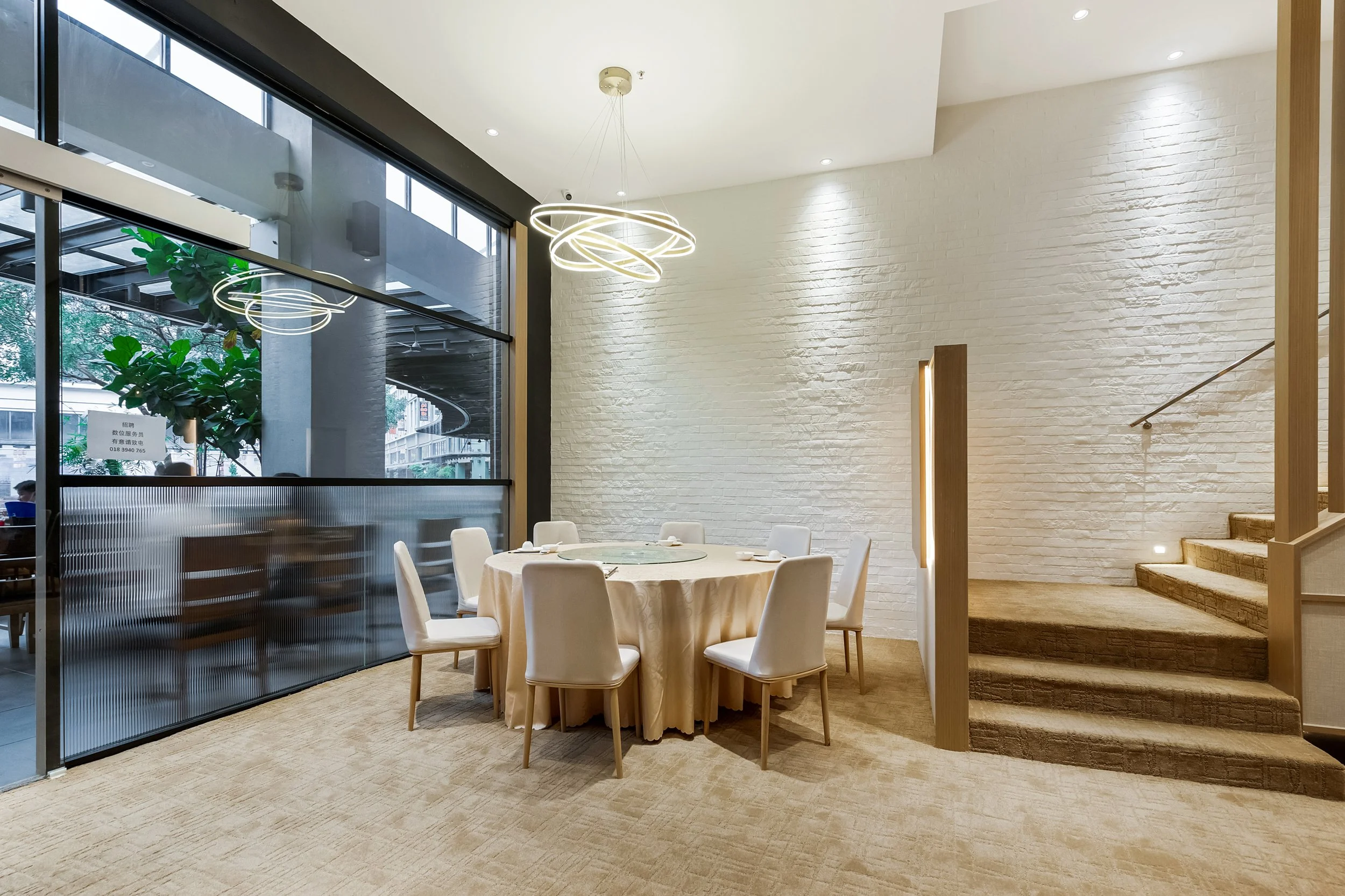 Empty round dining table with white chairs set with white tableware and a glass Lazy Susan, located in a modern restaurant with large window, white brick wall, contemporary ceiling light fixtures, and carpeted stairs.