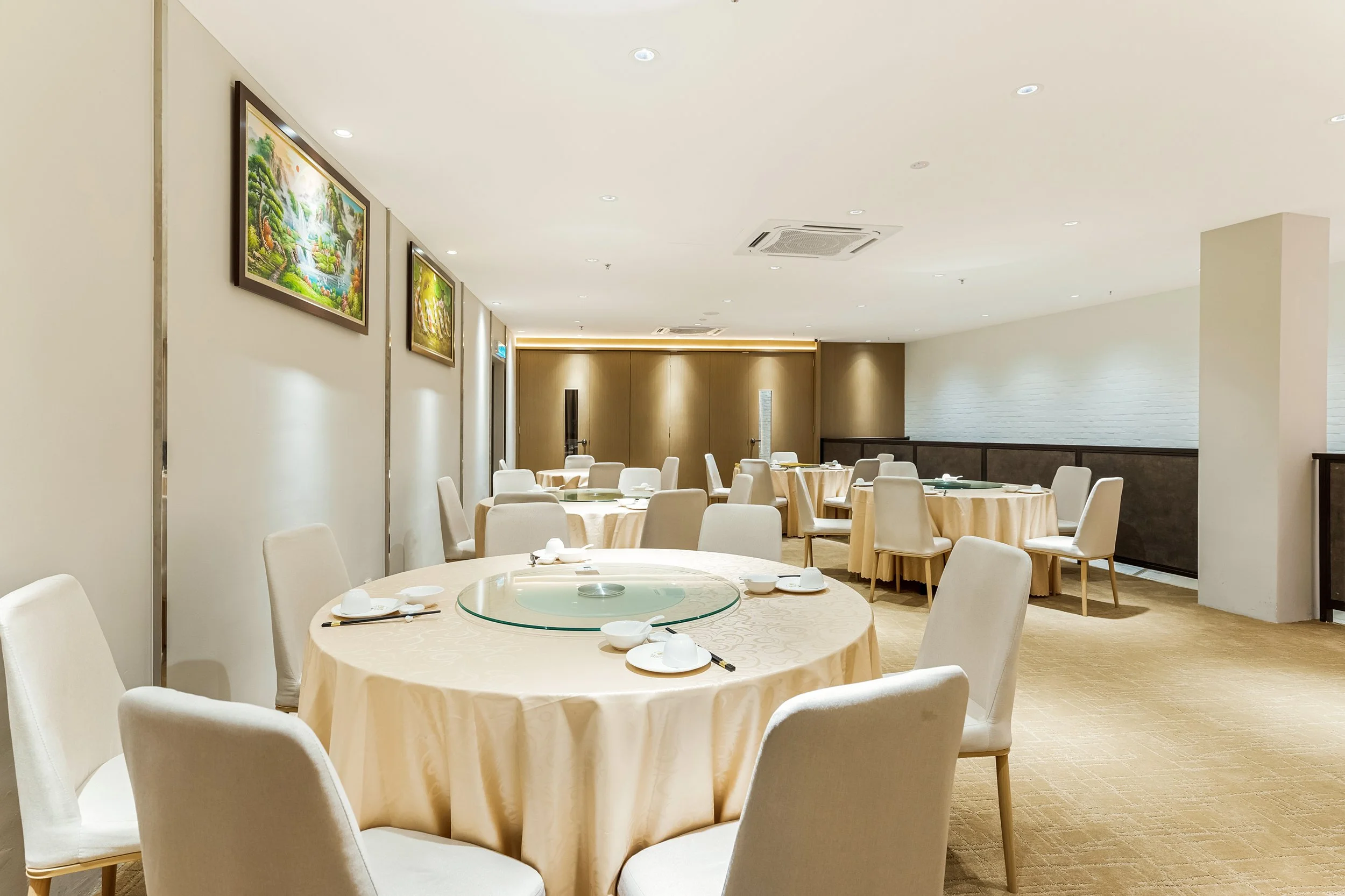 Empty dining room in a restaurant with round tables covered in beige tablecloths, set with white plates, bowls, chopsticks, and a glass lazy Susan at each table, beige chairs, paintings on the wall, and soft lighting.