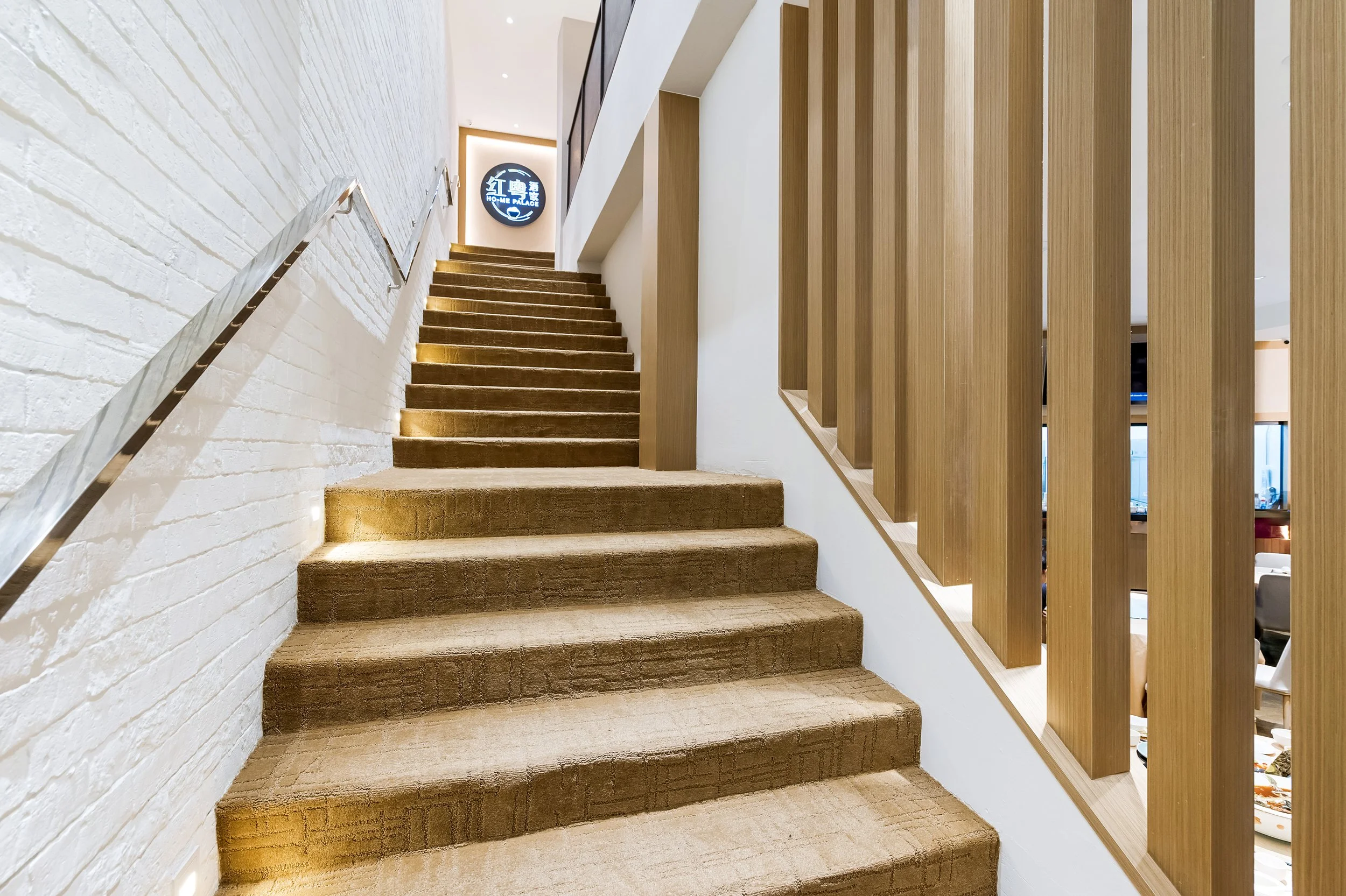 Indoor staircase with beige carpet, white brick wall on the left, wooden slats on the right, and a black sign with Asian characters and English text at the top of the stairs.