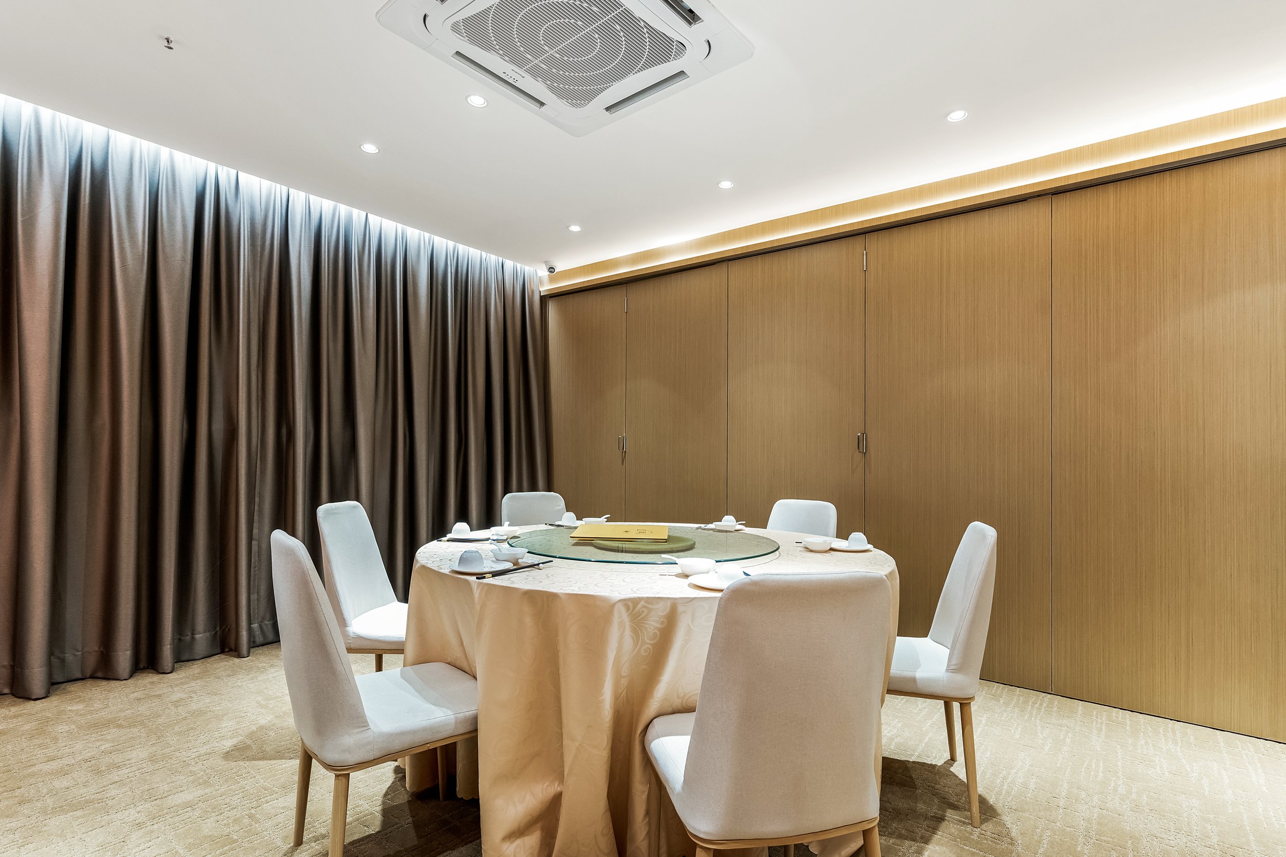 A round dining table set with white chairs, tableware, and a lazy Susan in a private dining room with beige carpeting, wood-paneled wall, and dark curtains.