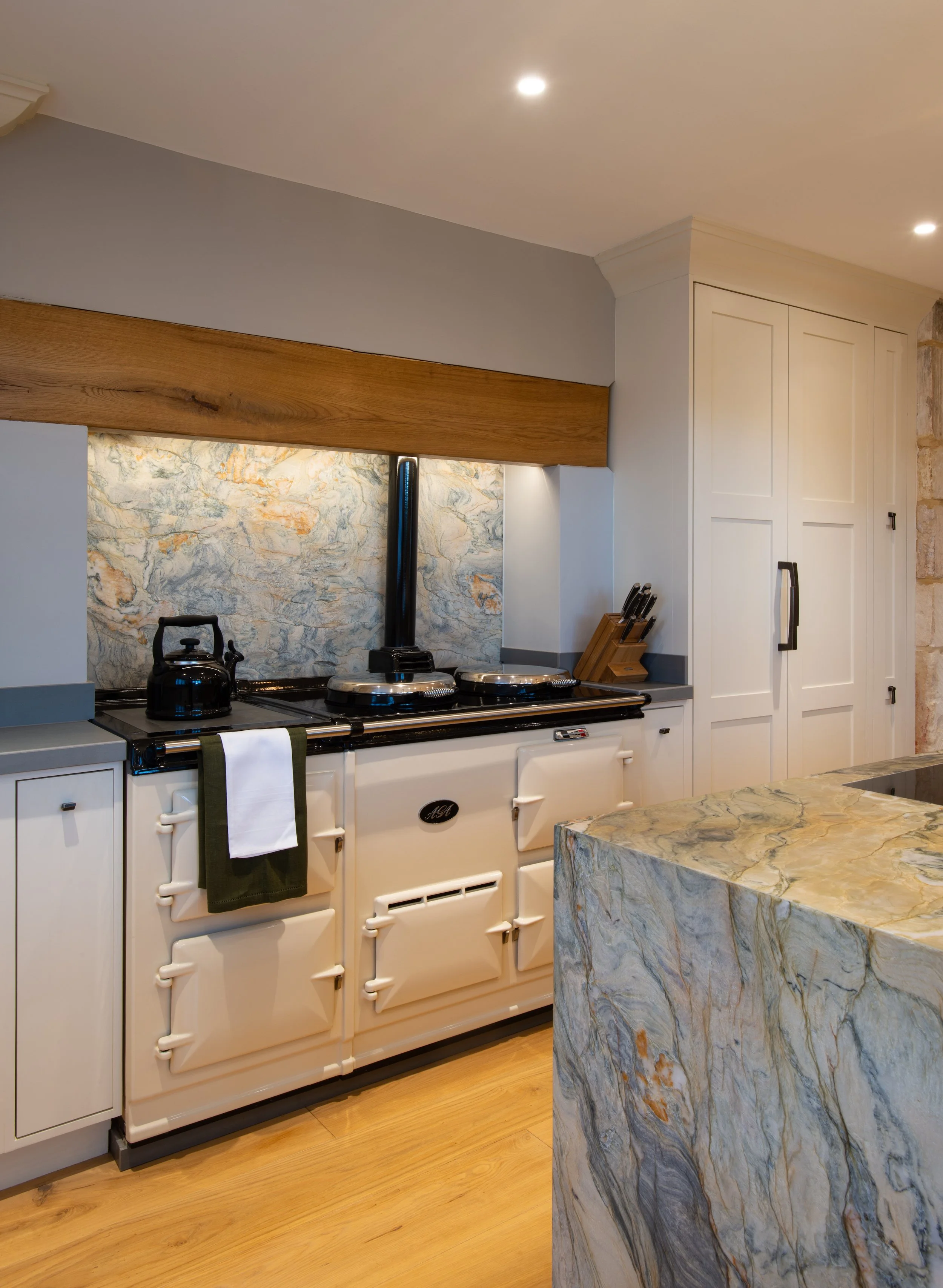 A detailed view of the cream AGA framed by bespoke painted cabinetry and a substantial oak beam. The strongly veined quartz used for the kitchen island is repeated as an AGA splashback, creating material continuity and a distinctive focal point withi