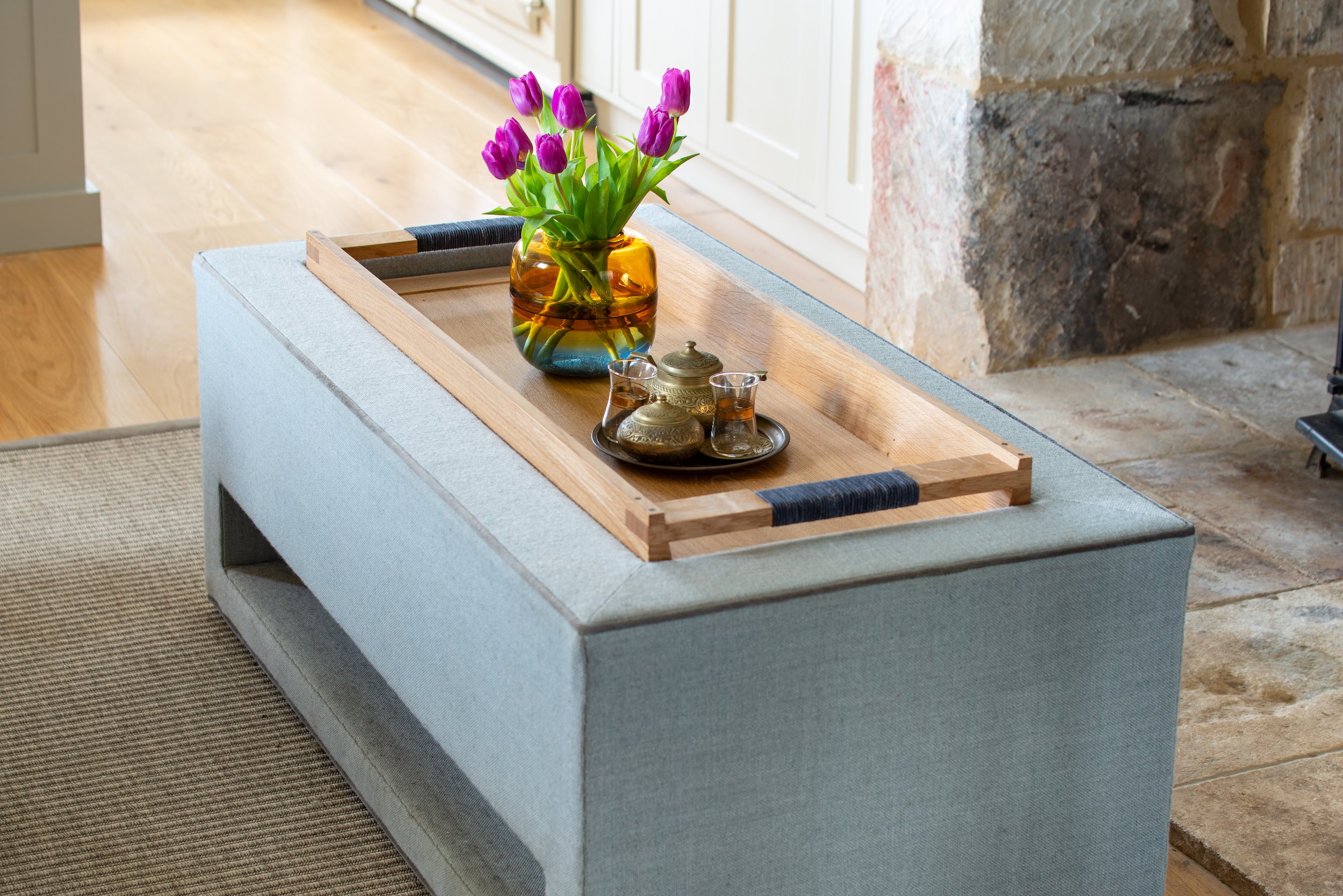 A detailed view of the bespoke upholstered ottoman, designed to function as both a coffee table and an integrated dog bed beside the stone fireplace. Hard-wearing indoor-outdoor fabric was selected to withstand dogs and muddy country clothing, while 