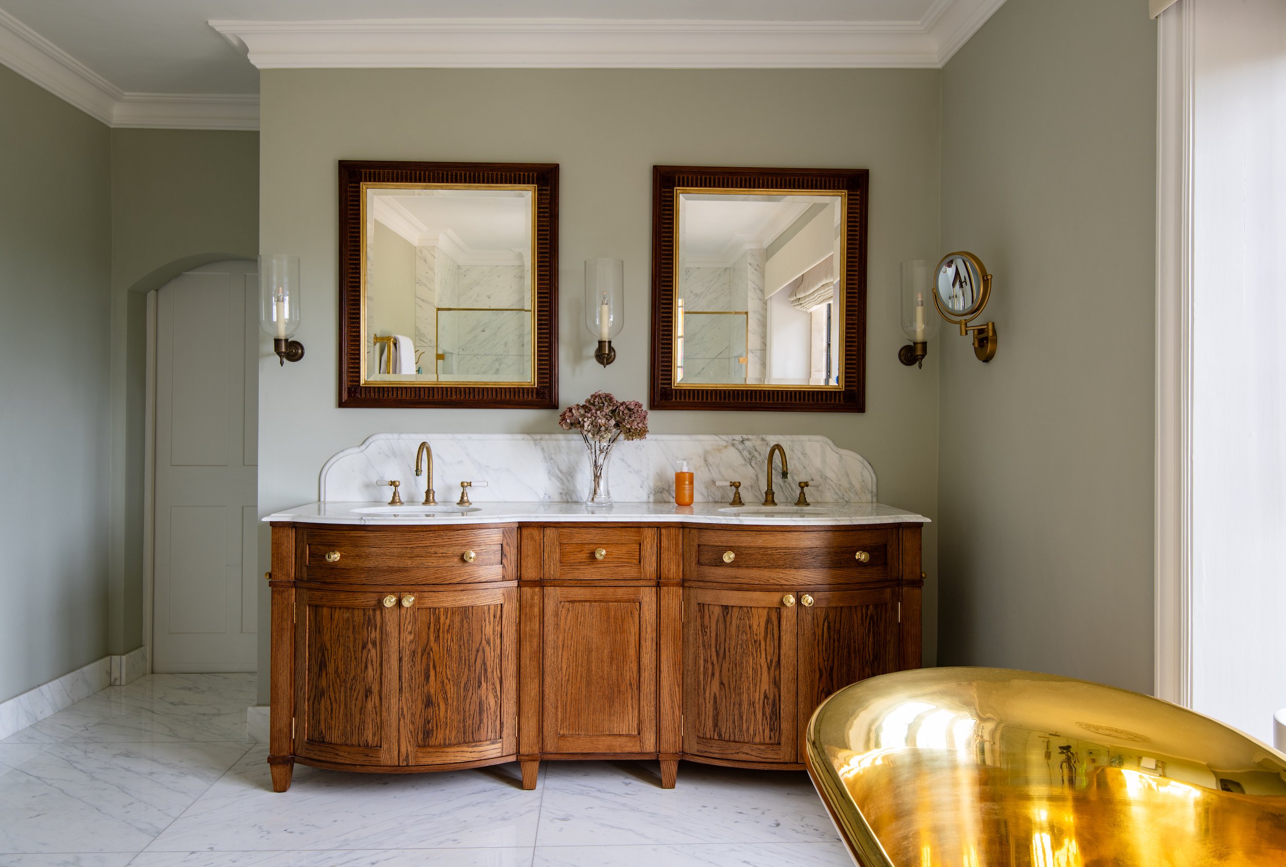 Master bathroom vanity designed by MIDA Interiors for a large Cotswolds country estate, featuring bespoke oak cabinetry, a shaped double marble basin top and splashback, antique-style mirrors, integrated lighting and brass fittings.