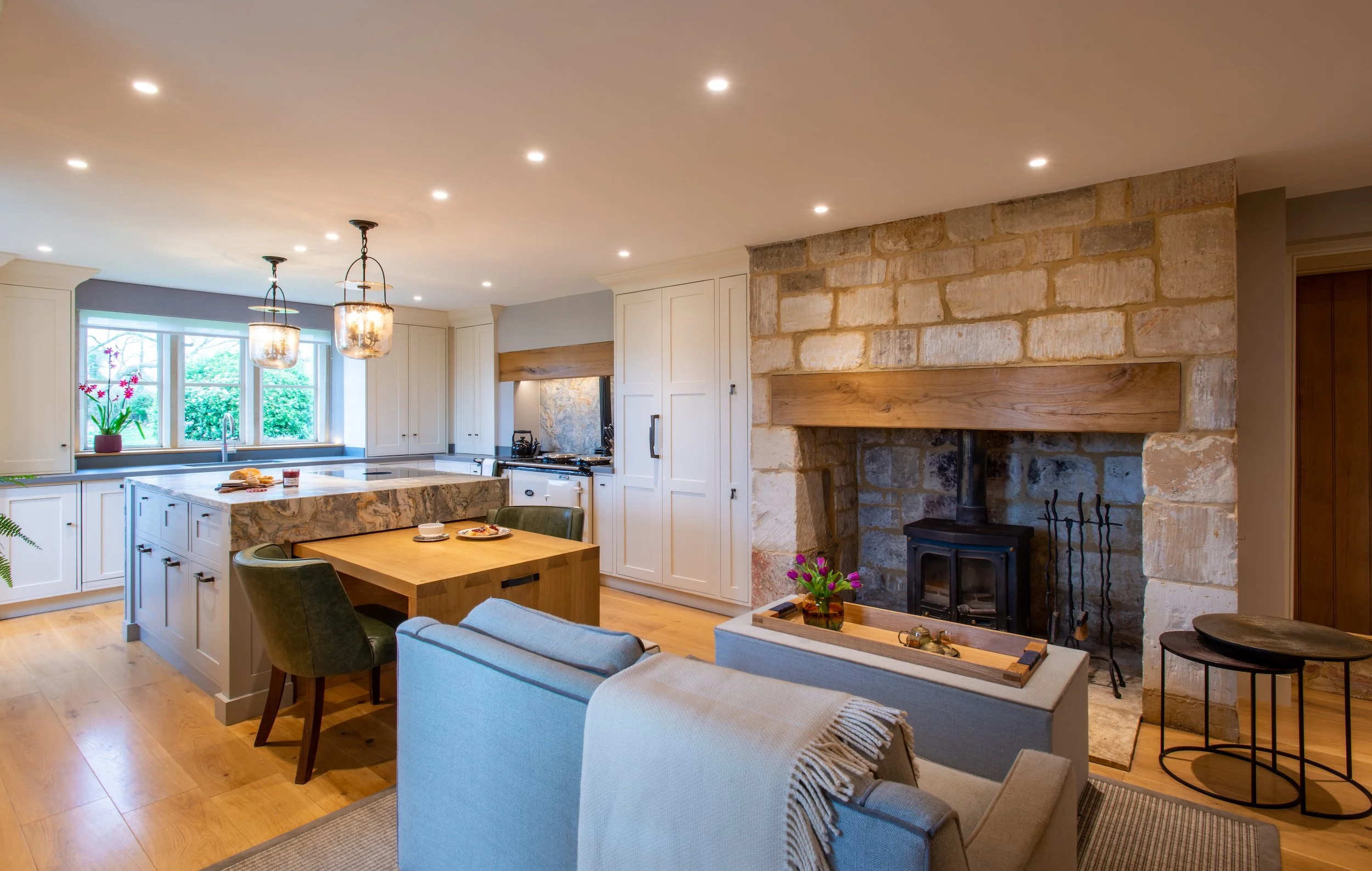 A view across the redesigned kitchen towards the substantial stone fireplace, showing the dovetail-jointed oak dining table extended from the bespoke island. The dual-purpose design provides a generous preparation surface and informal dining area whi