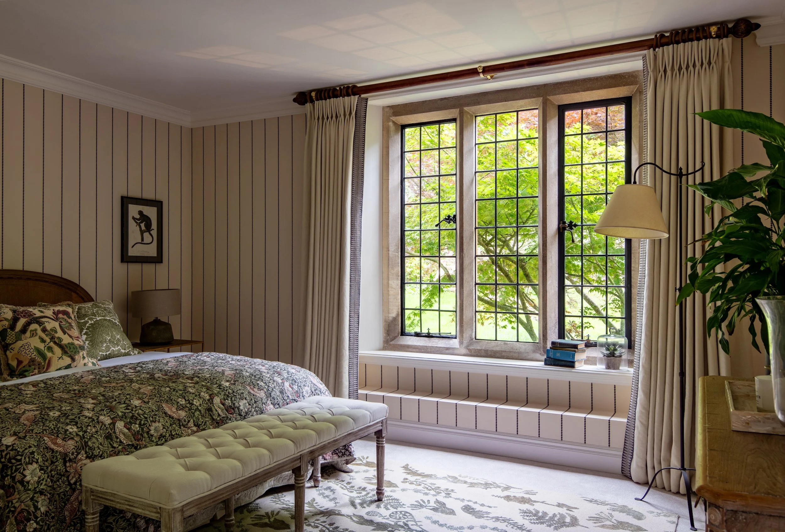 Guest bedroom designed by MIDA Interiors for a large Cotswolds country estate, featuring layered botanical textiles, tailored curtains, an upholstered bench, antique-inspired furniture and a calm palette of warm neutrals and soft greens.