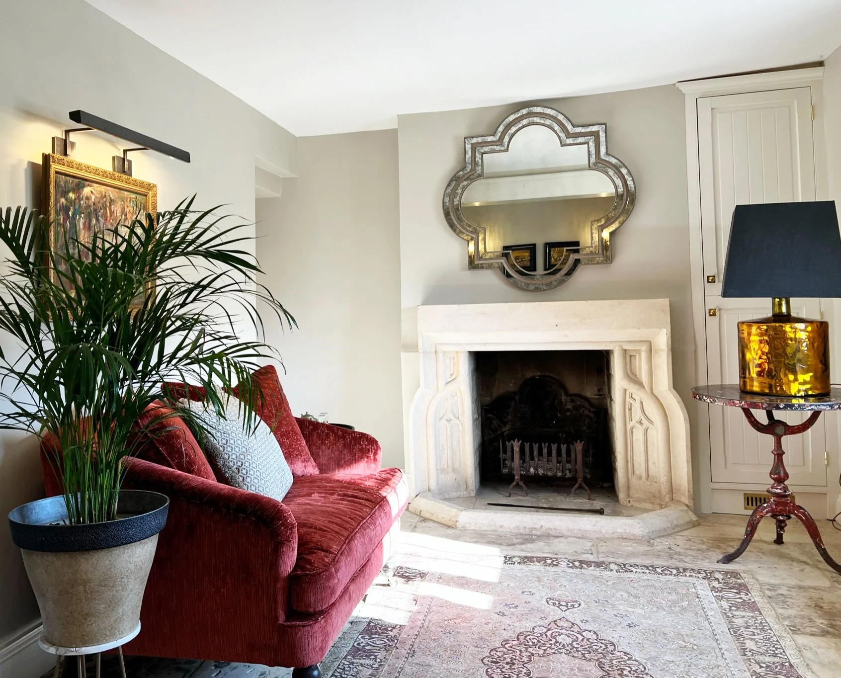 Entrance hall interior designed by MIDA Interiors for a Cotswolds country house, featuring a carved stone fireplace, original flag stone floor, red velvet sofa, decorative mirror, antique-style rug and bespoke fitted cabinetry.
