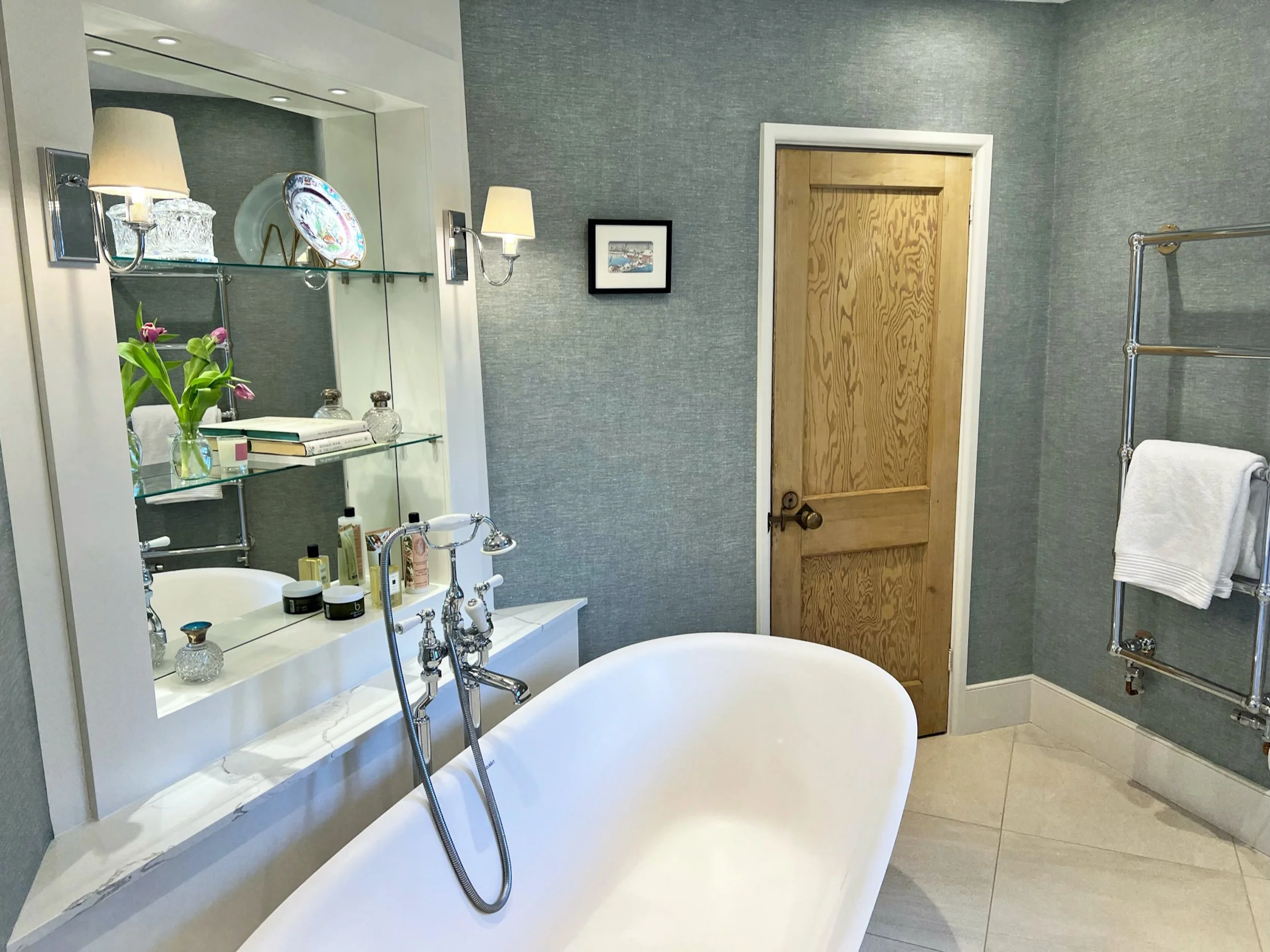 Freestanding bath framed by illuminated mirrored shelving, traditional polished fittings and textured linen look wallpaper in the family bathroom.