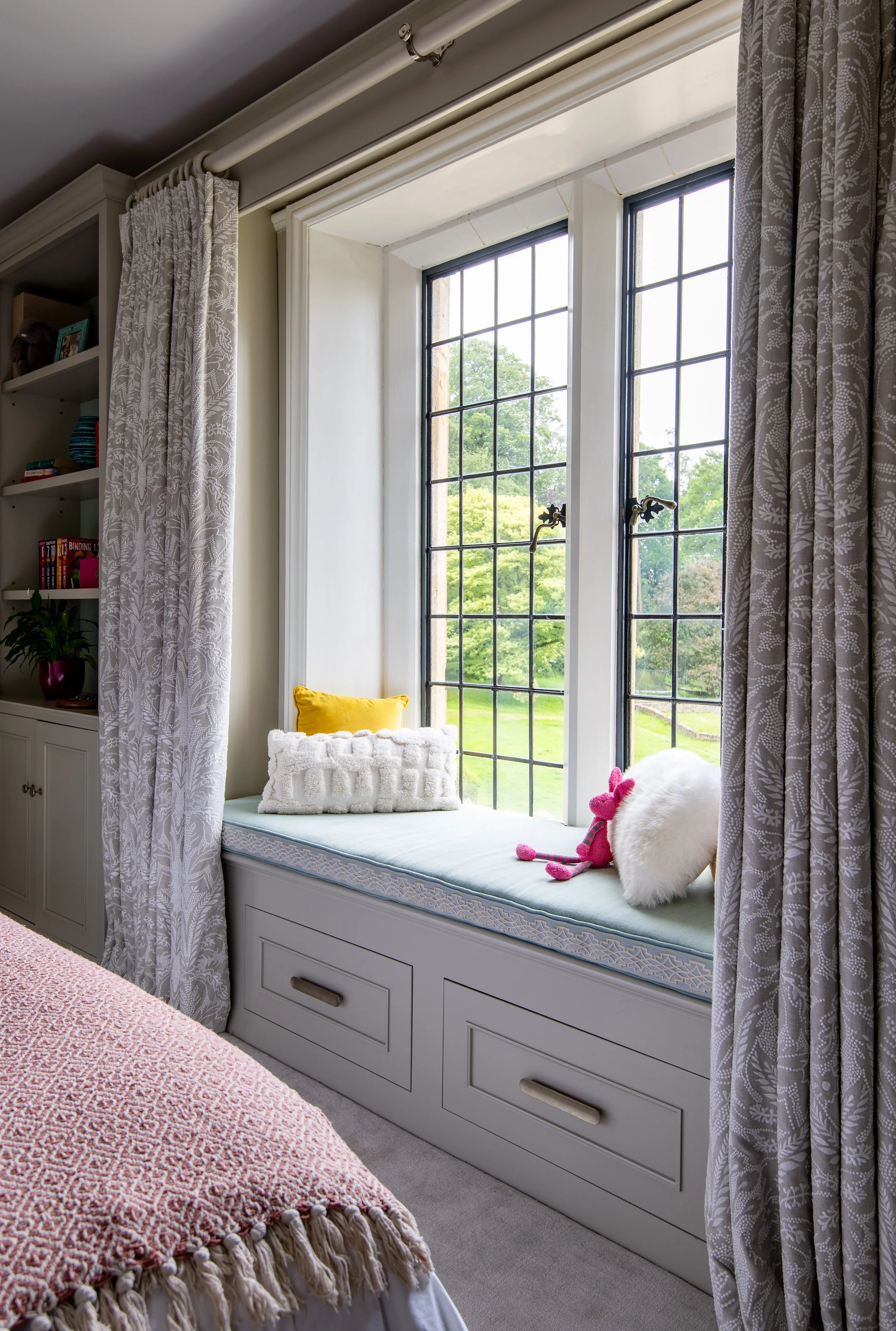 Children’s bedroom window seat designed by MIDA Interiors for a large Cotswolds country estate, featuring bespoke under-seat storage, tailored curtains and a cushioned reading nook overlooking the gardens.