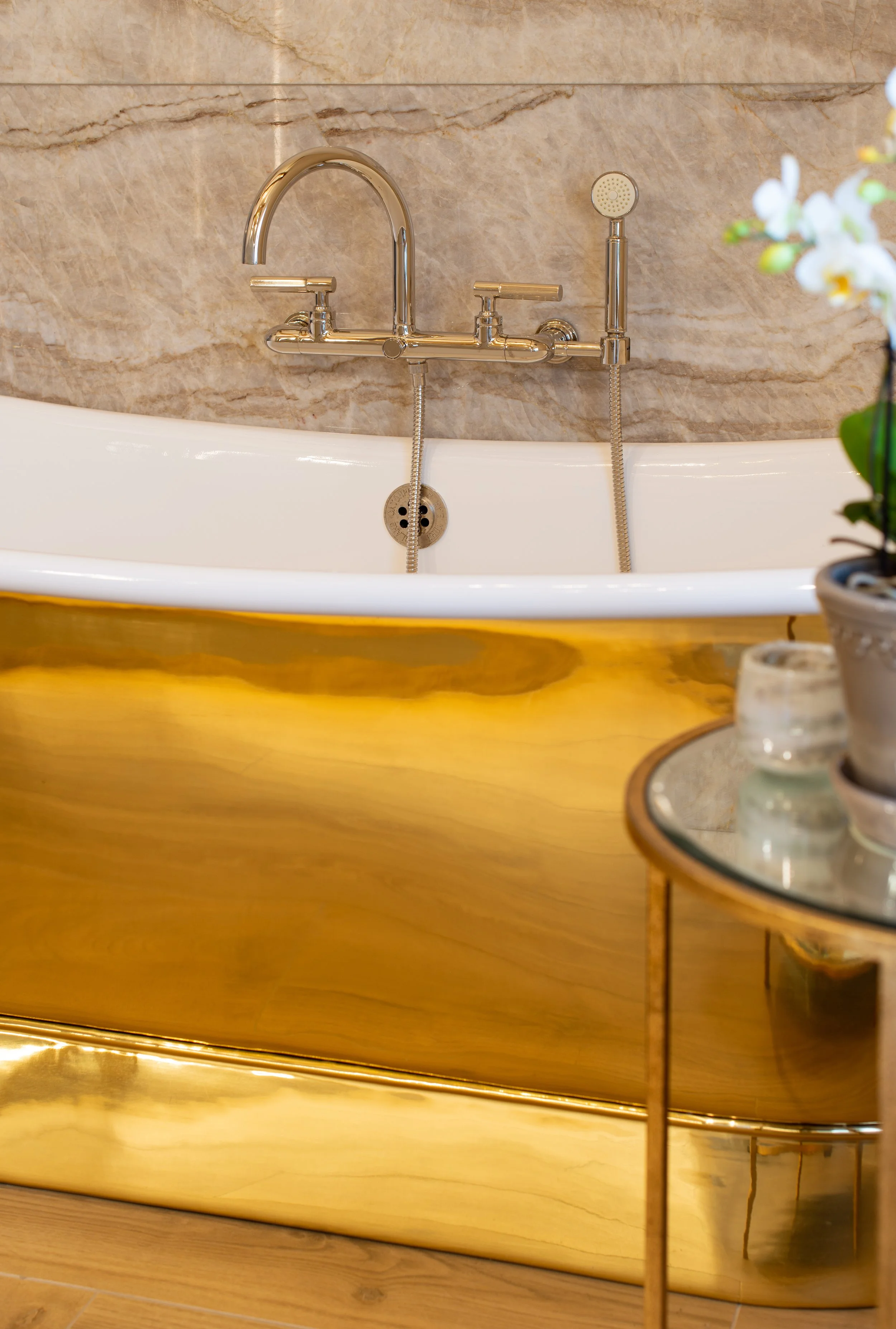 A detailed view of the deep freestanding soaking bath, finished with a polished brass exterior and traditional nickel bath fittings. The reflective metal introduces warmth against the softly veined stone backdrop, creating a distinctive focal point w