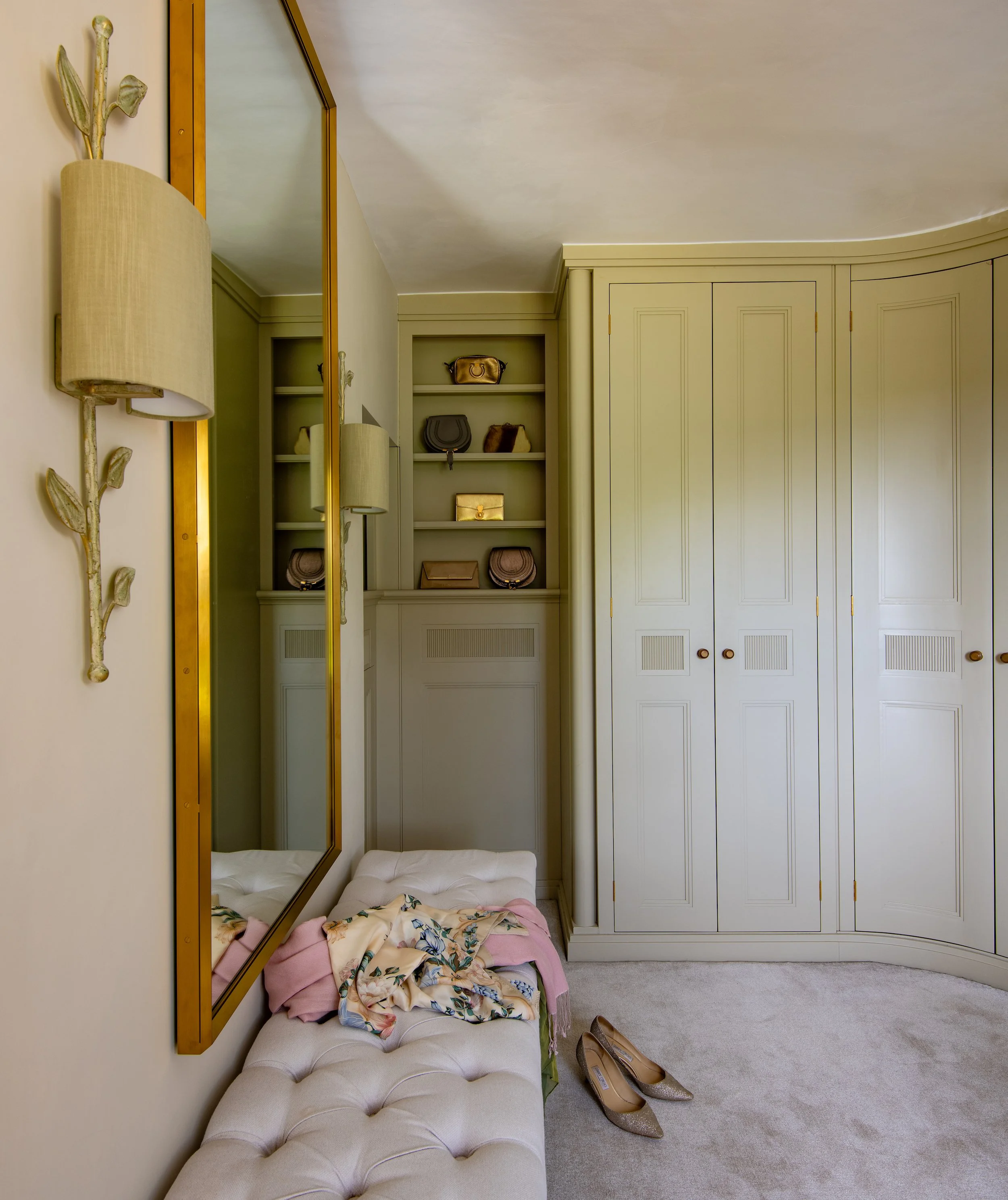 Master dressing room designed by MIDA Interiors for a large Cotswolds country estate, reconfigured from the former master bathroom with a curved wall, bespoke fitted wardrobes, integrated display shelving, a full-height mirror and tailored lighting.