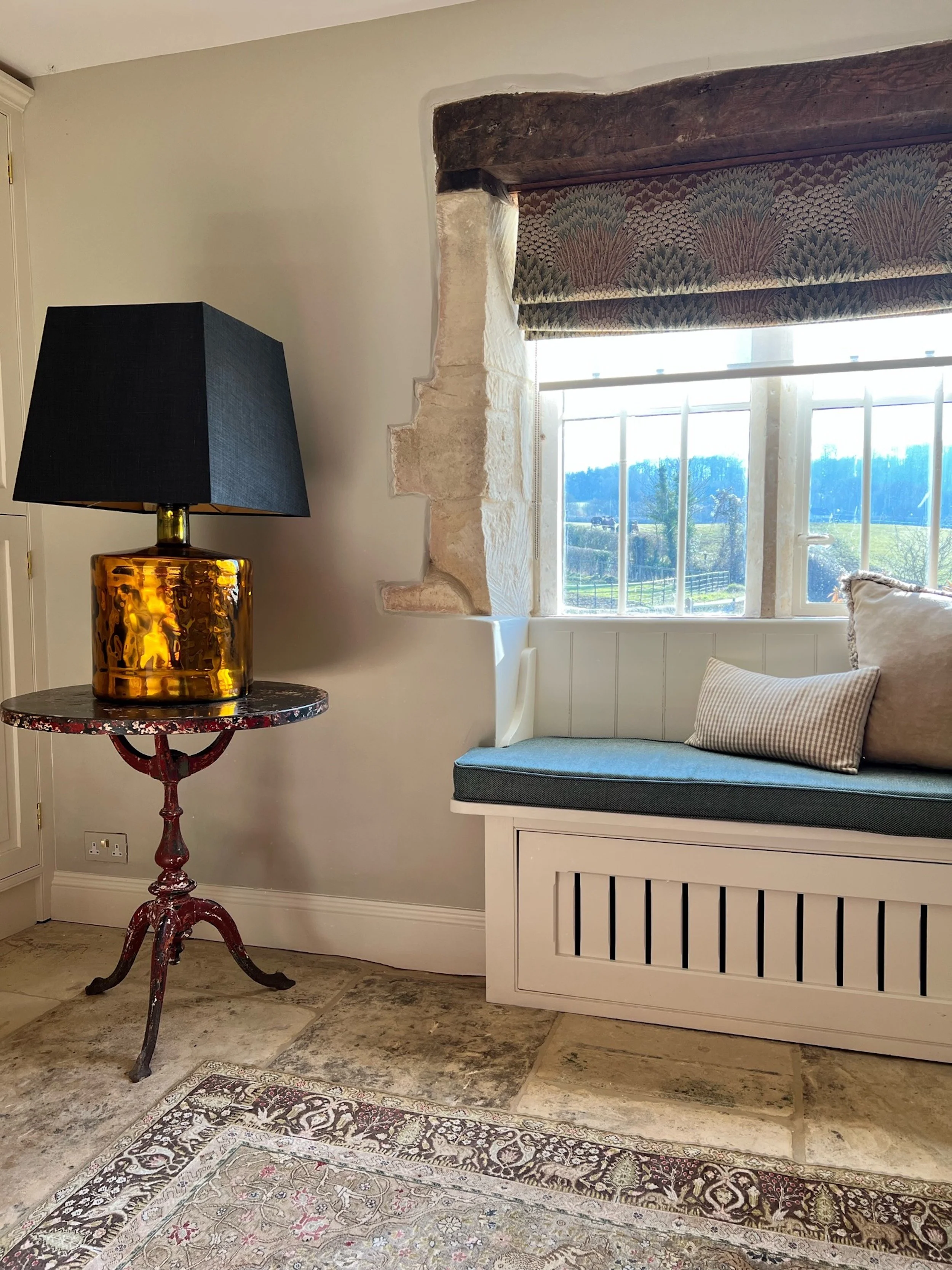 A bespoke window seat makes considered use of the deep stone-framed window within the entrance hall, creating a quiet place from which to enjoy views across the surrounding countryside. Tailored upholstery, a patterned Roman blind and an amber glass 
