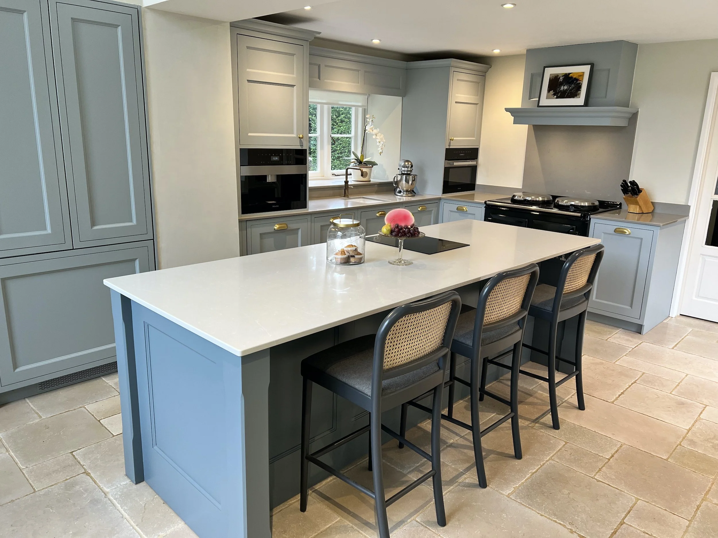 Painted Shaker kitchen in a 19th-century Oxfordshire cottage, with a stone-topped island, cane backed seating, integrated appliances and traditional stone flooring.