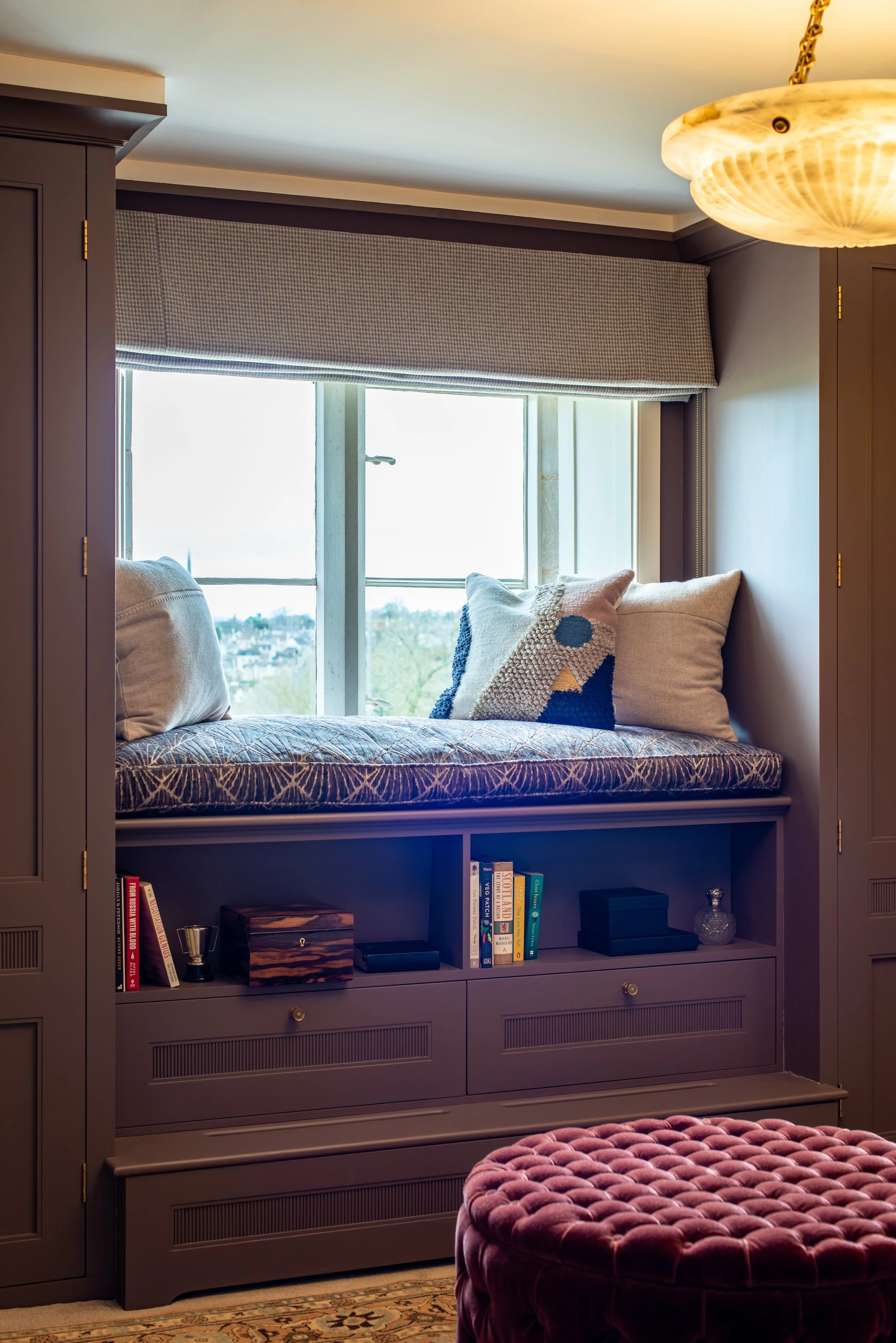 A bespoke upholstered window seat is integrated between the full-height wardrobes in the gentleman’s dressing room. Open shelving and reeded drawers provide additional storage below, while a tailored Roman blind and layered cushions create a quiet pl