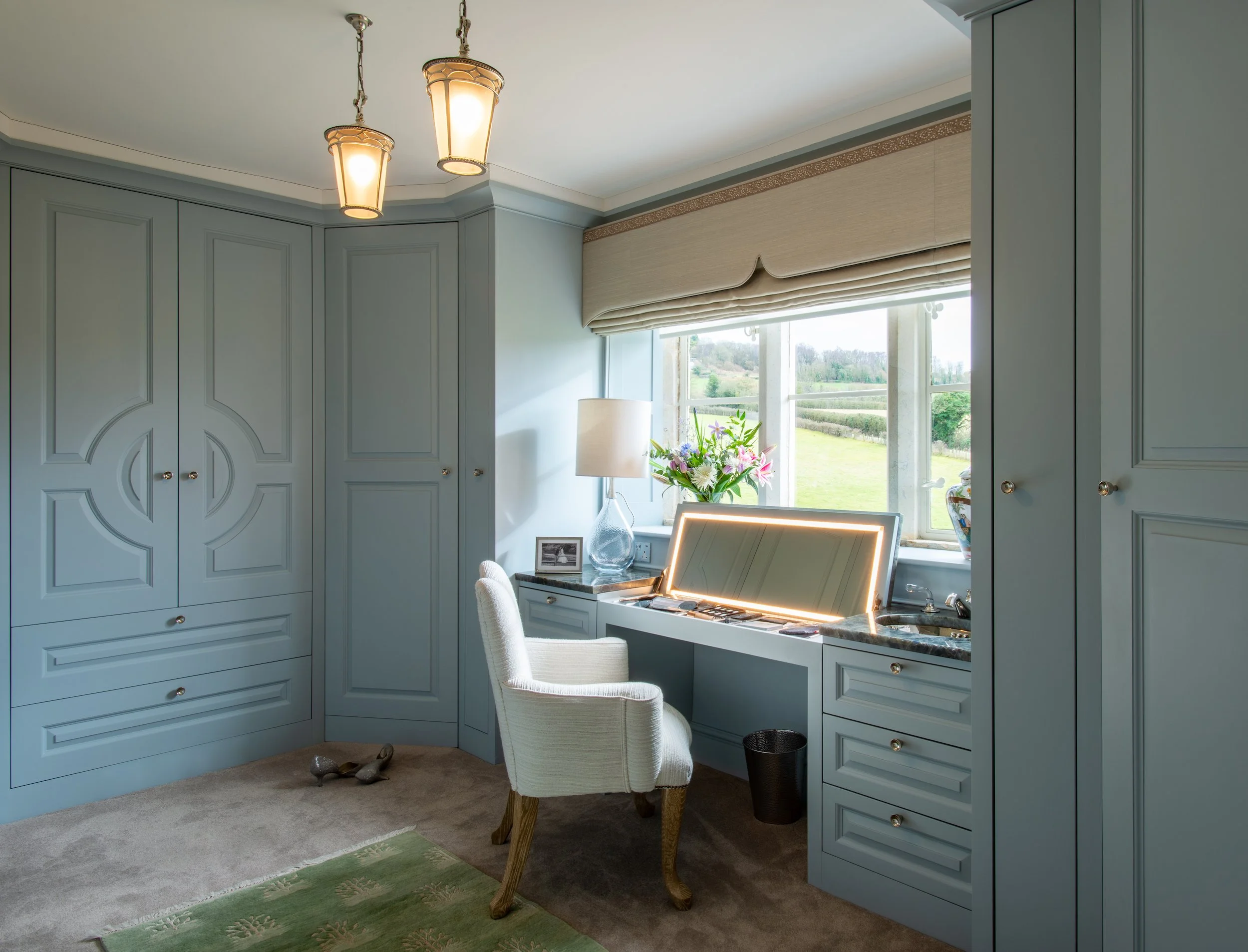 The second dressing room is arranged around pale blue bespoke cabinetry, with full-height wardrobes, integrated drawers and an illuminated lift-up vanity. Decorative door detailing, tailored pendant lights and a shaped Roman blind introduce character
