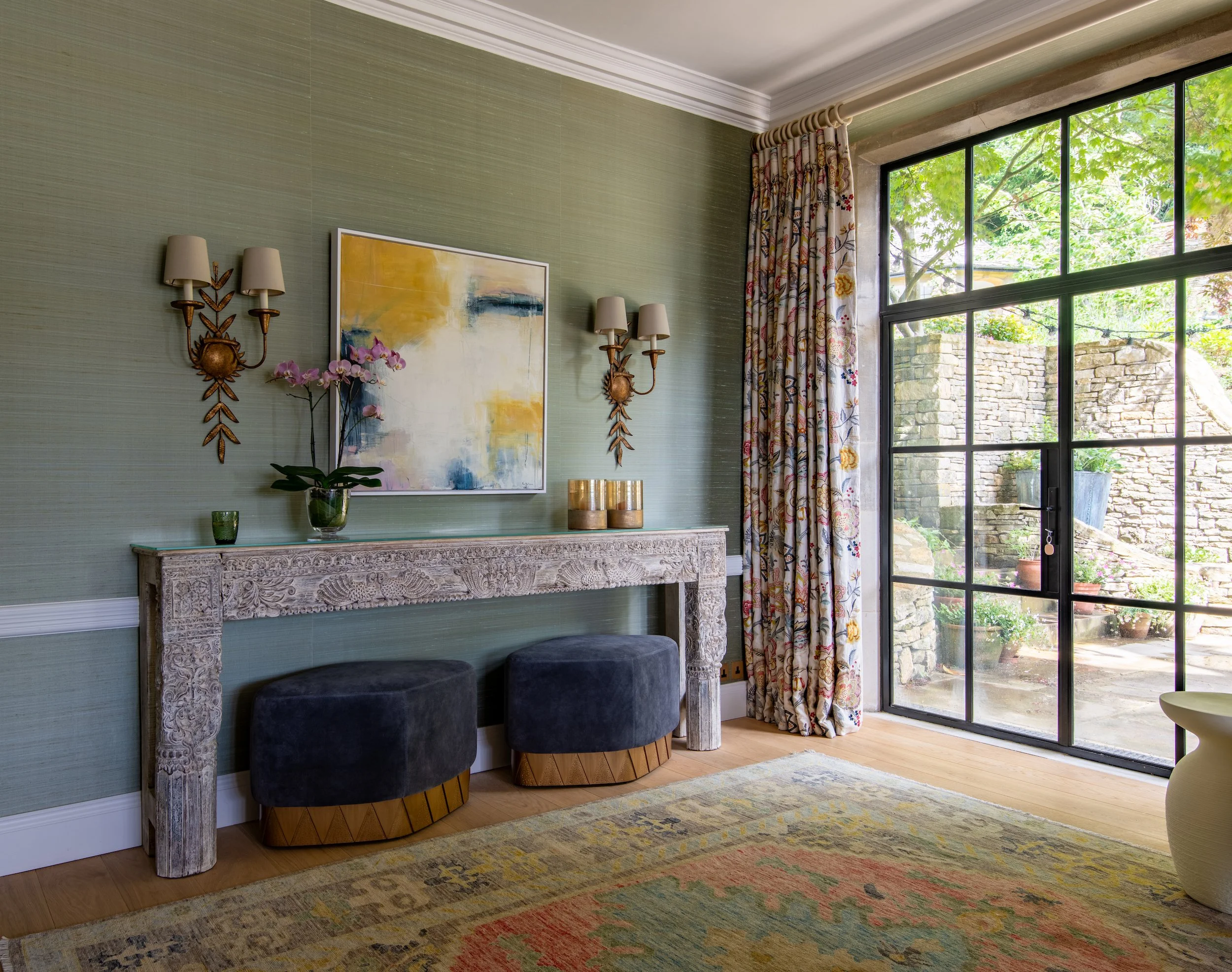Drawing room designed by MIDA Interiors for a large Cotswolds country estate, featuring silk walling, curated artwork, a carved console table, upholstered ottomans and Crittall French doors opening onto the garden terrace.
