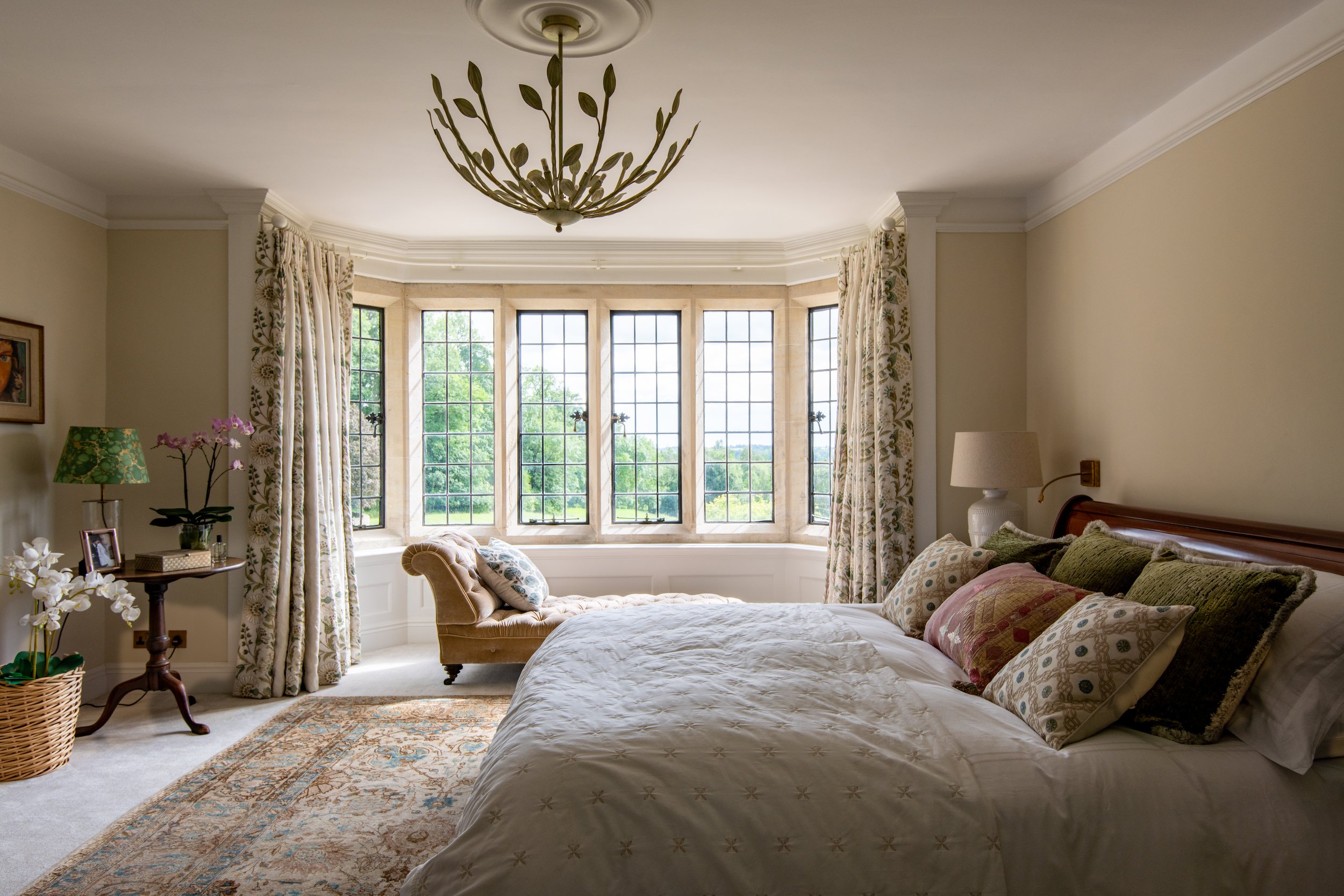 Master bedroom designed by MIDA Interiors for a large Cotswolds country estate, featuring a timber sleigh bed, layered bespoke soft furnishings, an antique rug, tailored curtains and a bay-window chaise overlooking the grounds.