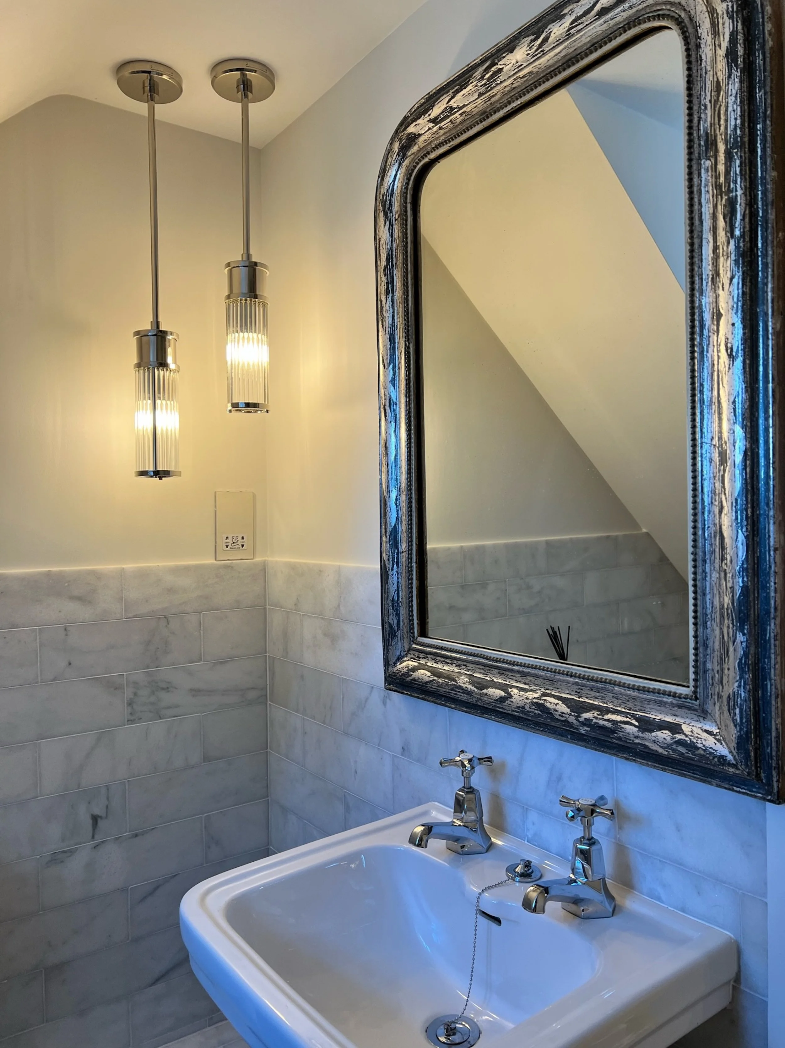 A traditional ceramic basin and crosshead taps are paired with a generously scaled, distressed-frame mirror in the guest bathroom. Contemporary fluted pendant lights bring warmth and vertical emphasis to the compact space, while pale marble wall tile