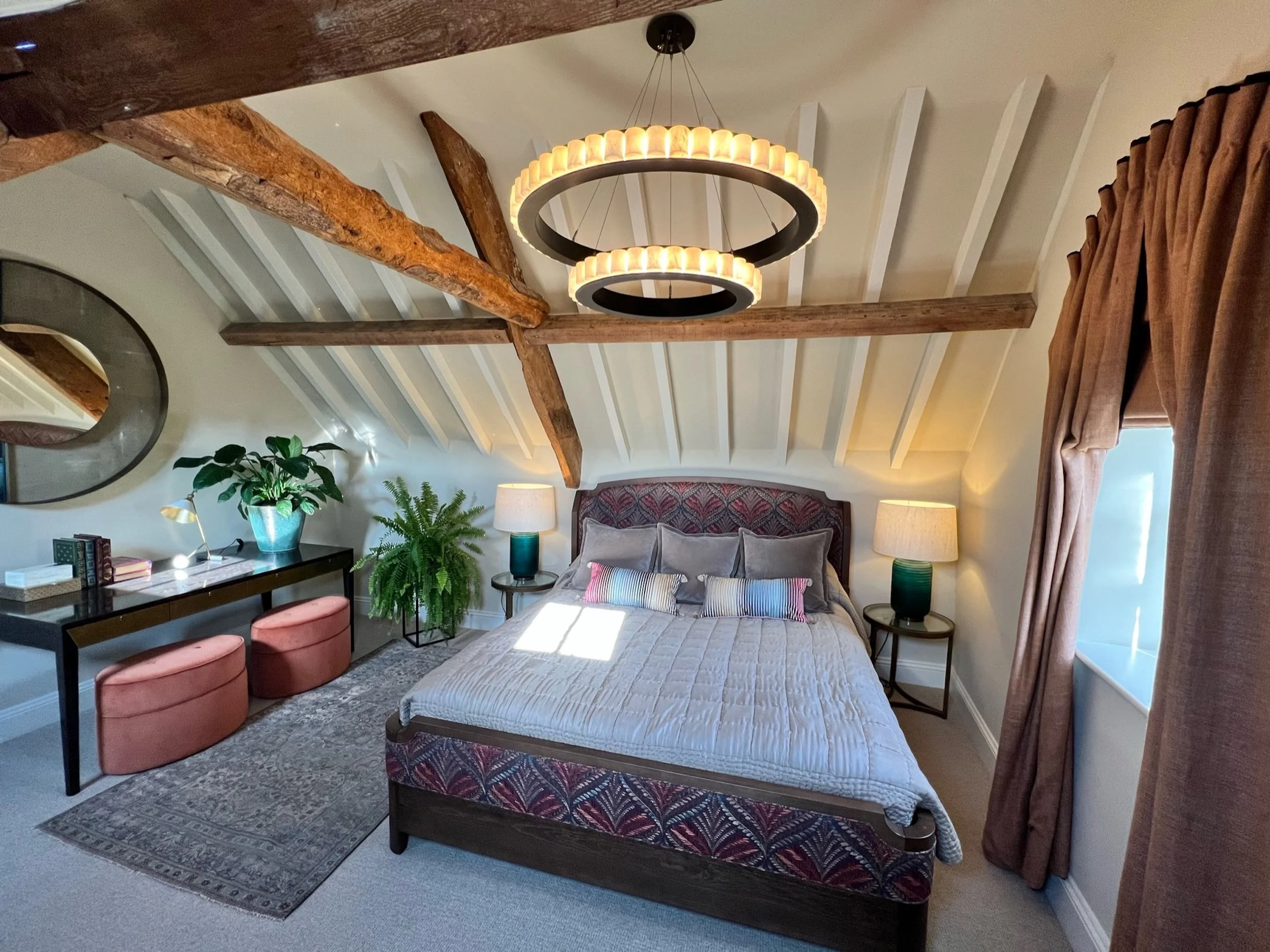 A wide view of the principal guest bedroom reveals the full volume of its vaulted ceiling and exposed historic timber structure. Contemporary tiered pendant lighting draws attention to the height of the room, while a richly upholstered bed, tailored 