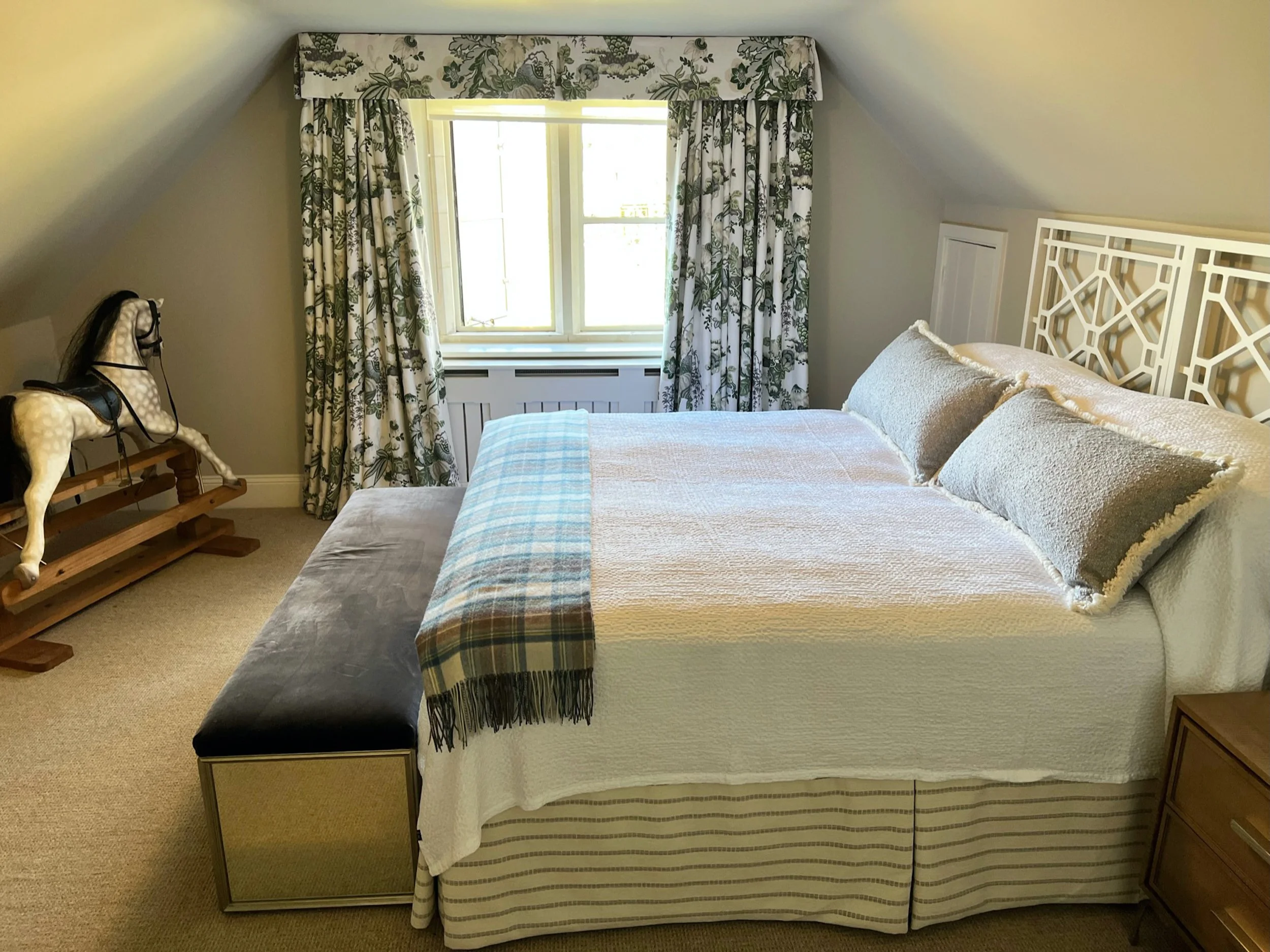 A flexible twin guest bedroom within the Cotswold country house, designed beneath the room’s sloping roofline. Bespoke geometric headboards and full-length botanical curtains introduce structure and pattern, while softly textured bedding and a checke
