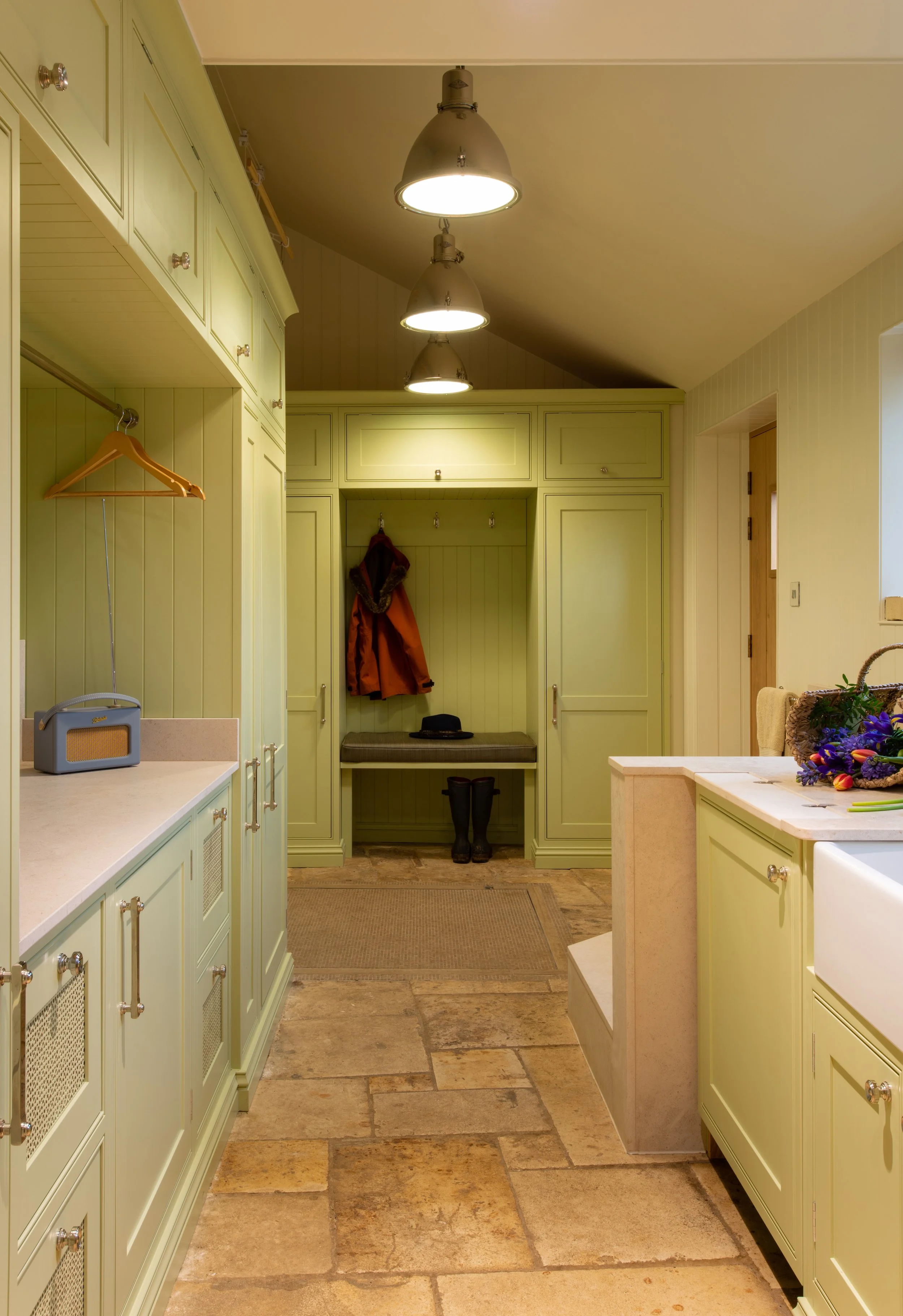 A combined boot room, utility and dog-wash area within a Grade II listed Cotswold farmhouse. Sage-green bespoke joinery provides extensive storage while concealing an electric drying cupboard and integrated commercial-grade laundry appliances. A leve