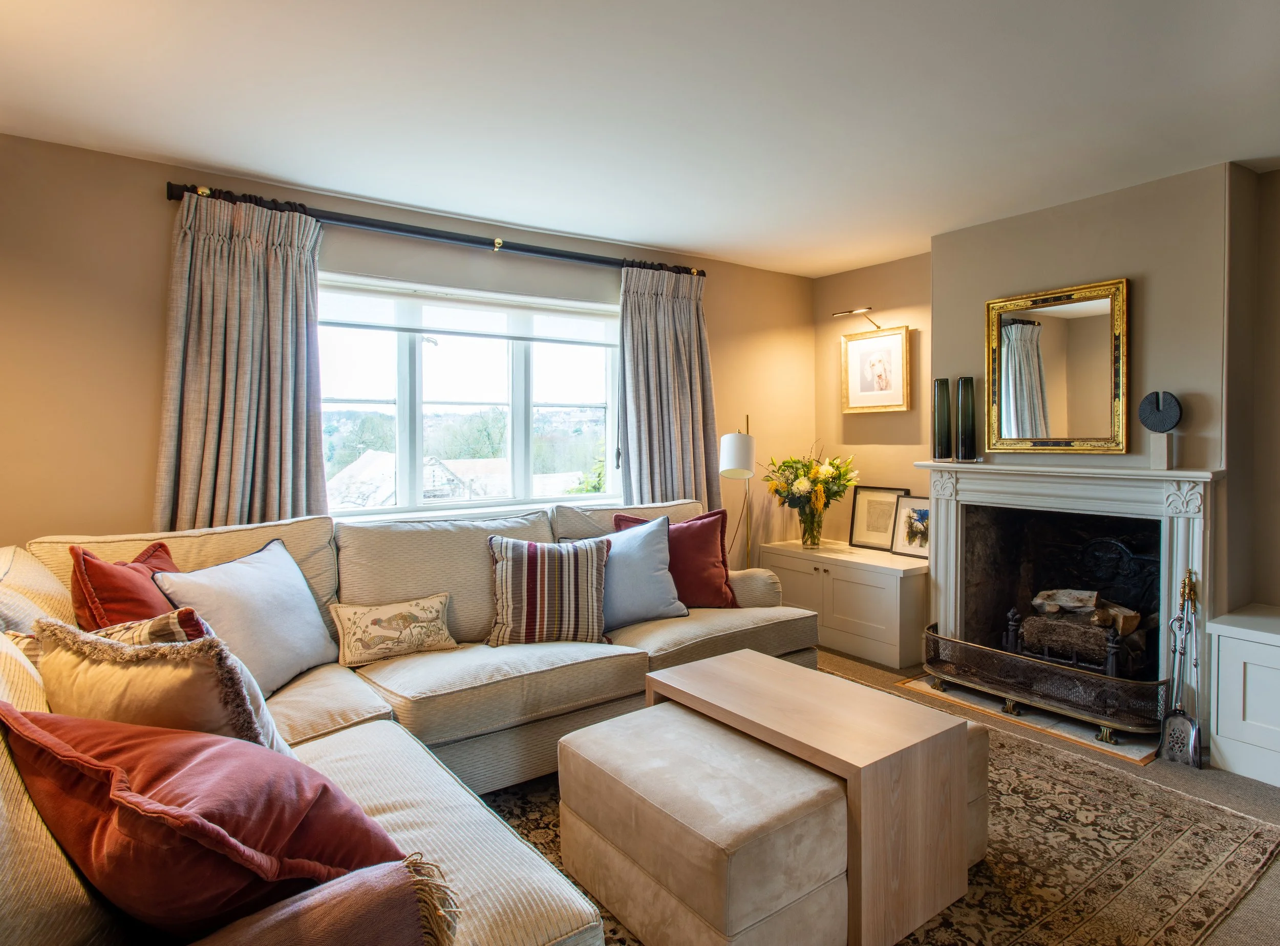 An inviting living room adjoining the library and games room within a Grade II listed Cotswold farmhouse. A generous L-shaped sofa and upholstered ottoman create flexible seating for informal entertaining, arranged around an oak coffee table and trad