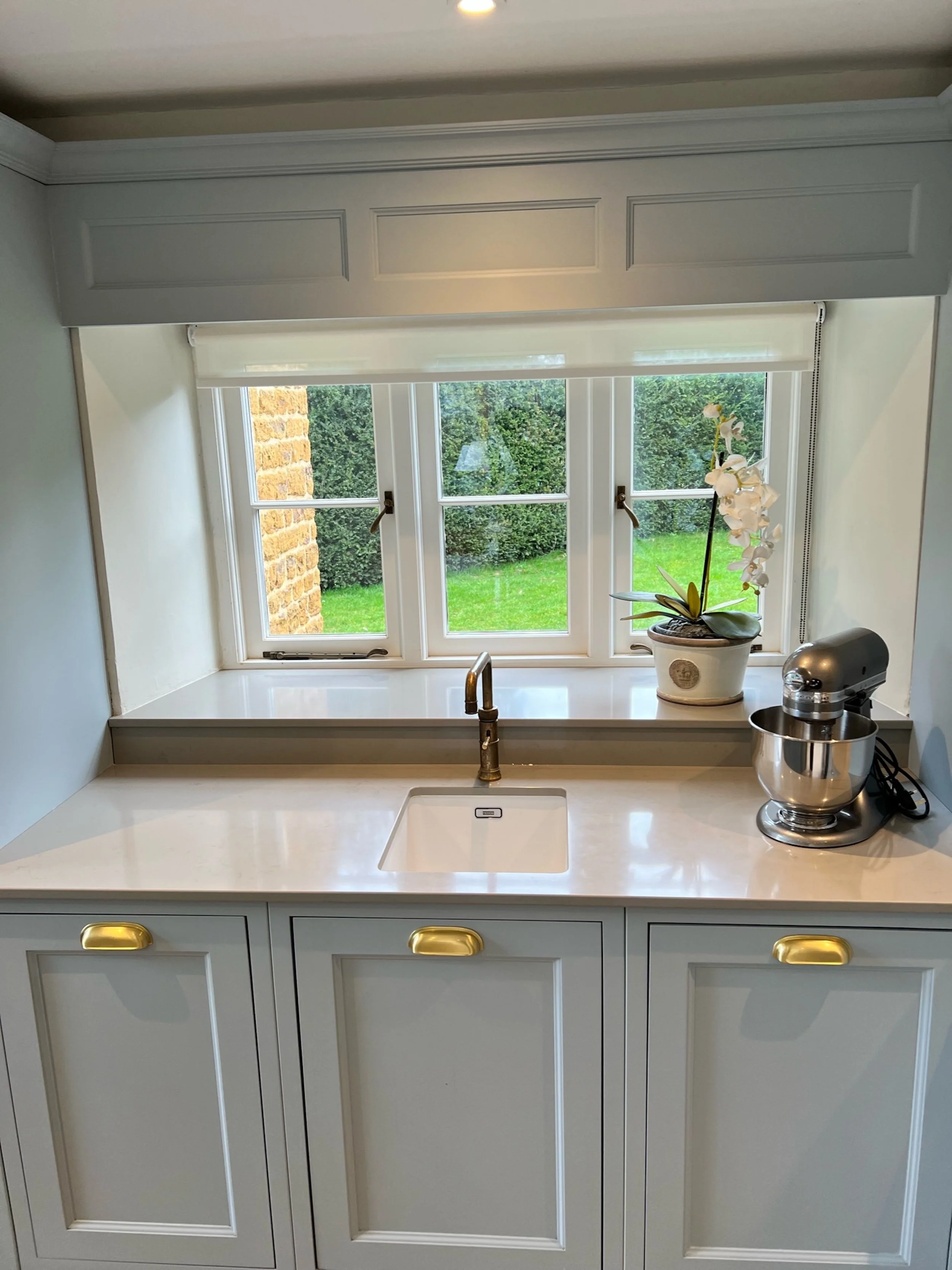 Under mounted kitchen sink overlooking the cottage garden, framed by painted cabinetry, brass hardware and a pale stone worktop.