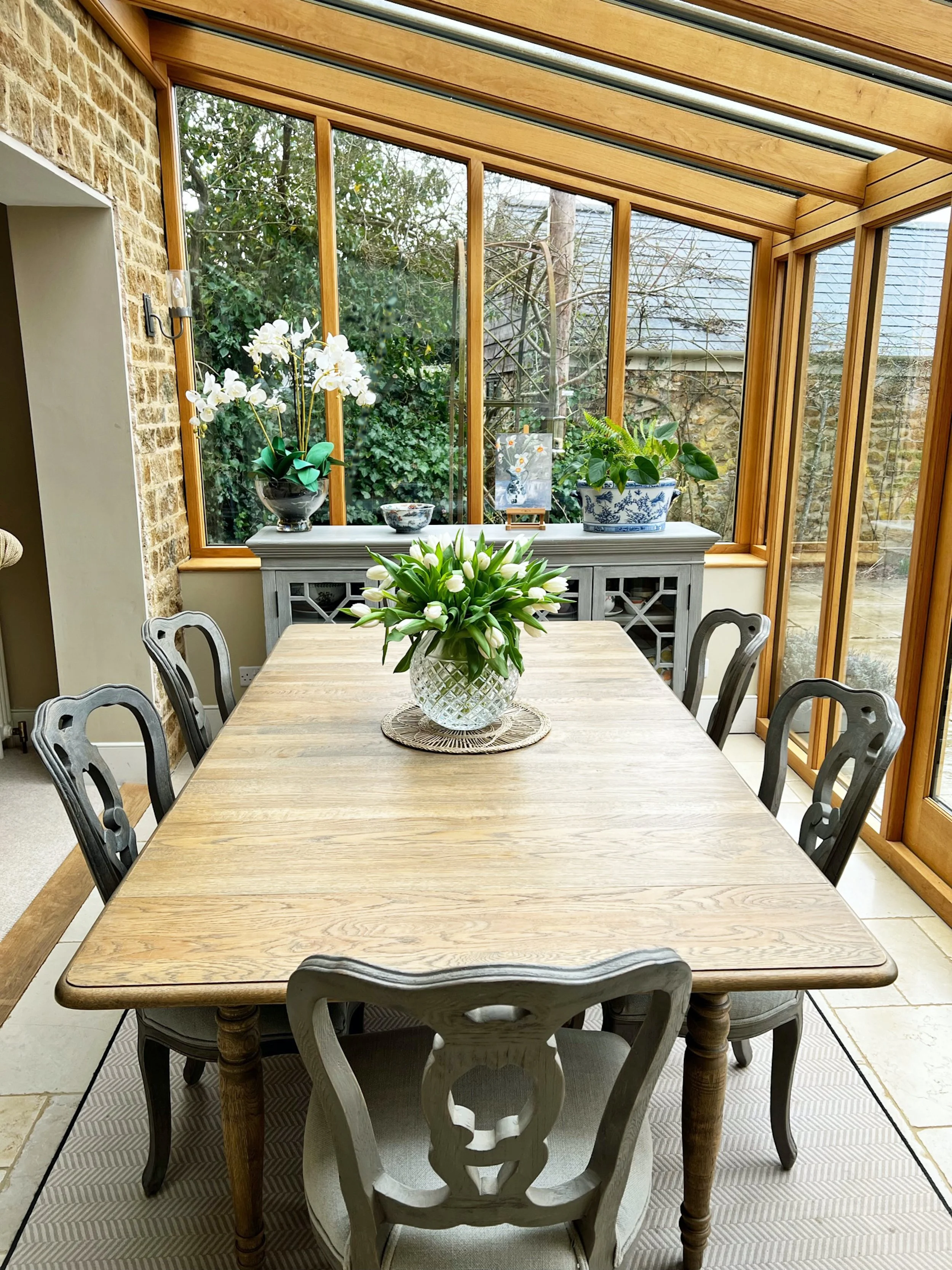 Country dining table and painted chairs set beneath a glazed timber roof, complemented by a decorative sideboard, natural textures and garden views.