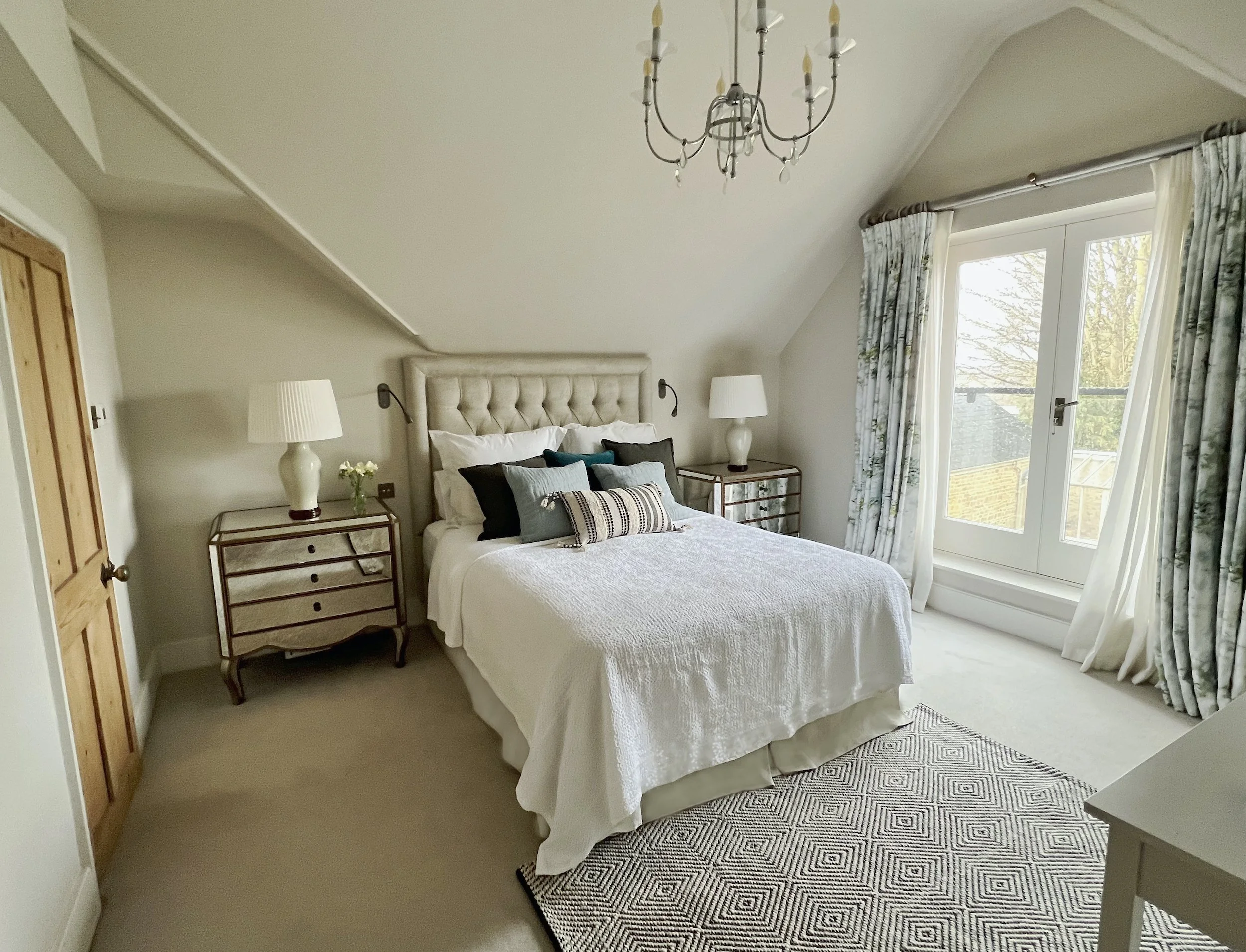 Principal bedroom in a 19th-century Oxfordshire cottage, with an upholstered bed, layered textiles, mirrored bedside chests and bespoke curtains framing a Juliet balcony and garden views.