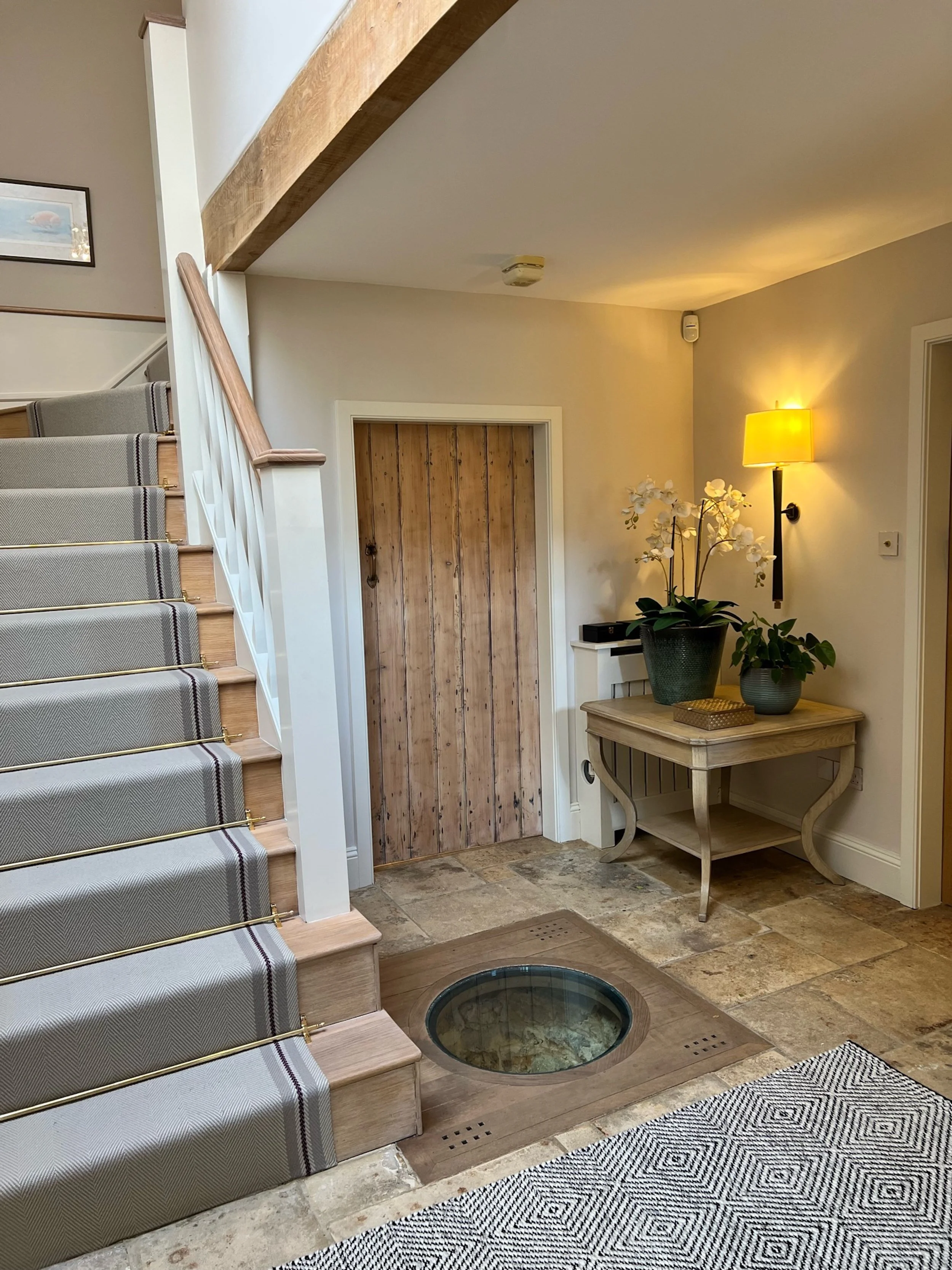 The stair hall brings together retained architectural features and carefully introduced contemporary elements, including a glazed viewing panel set into the floor above the historic well. A tailored stair runner, exposed oak beam, reclaimed timber do