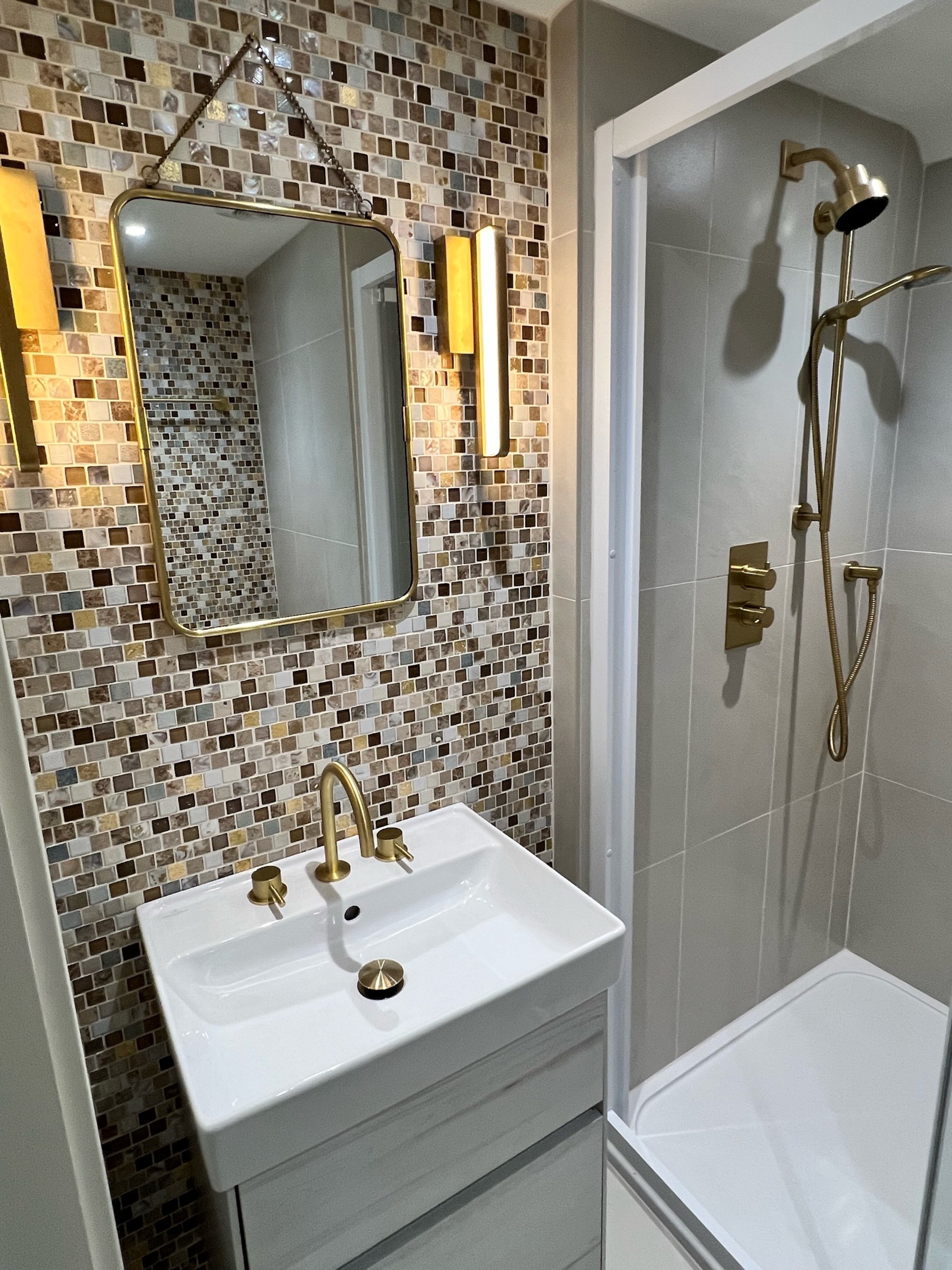 The compact shower room adjoining the principal guest bedroom is carefully planned to balance function with material interest. A mosaic tiled feature wall frames the brass-edged mirror and contemporary wall lights, while brushed brass fittings introd