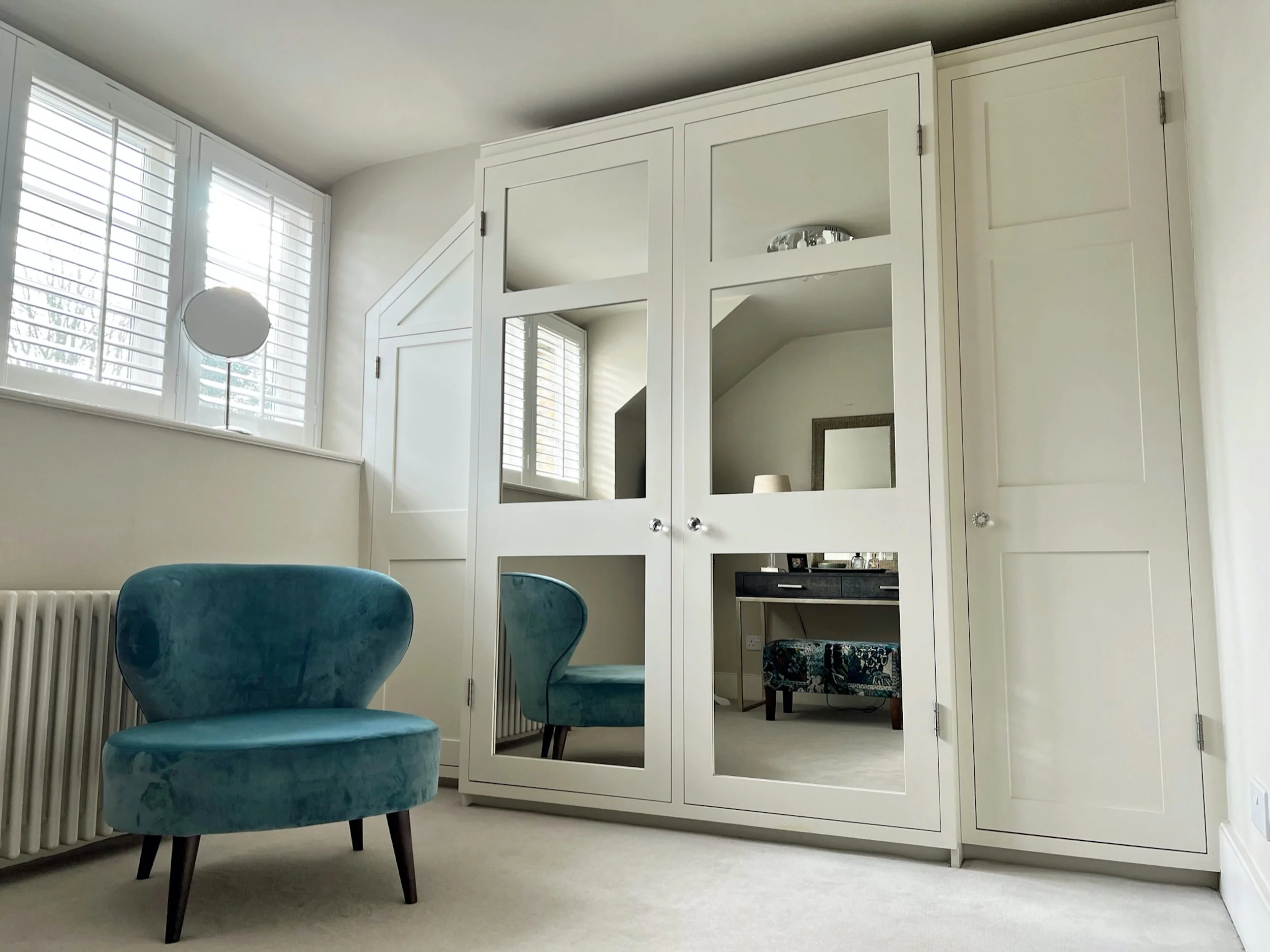 Tailored dressing room with floor-to-ceiling fitted wardrobes, mirrored doors, plantation shutters and a sculptural teal occasional chair.